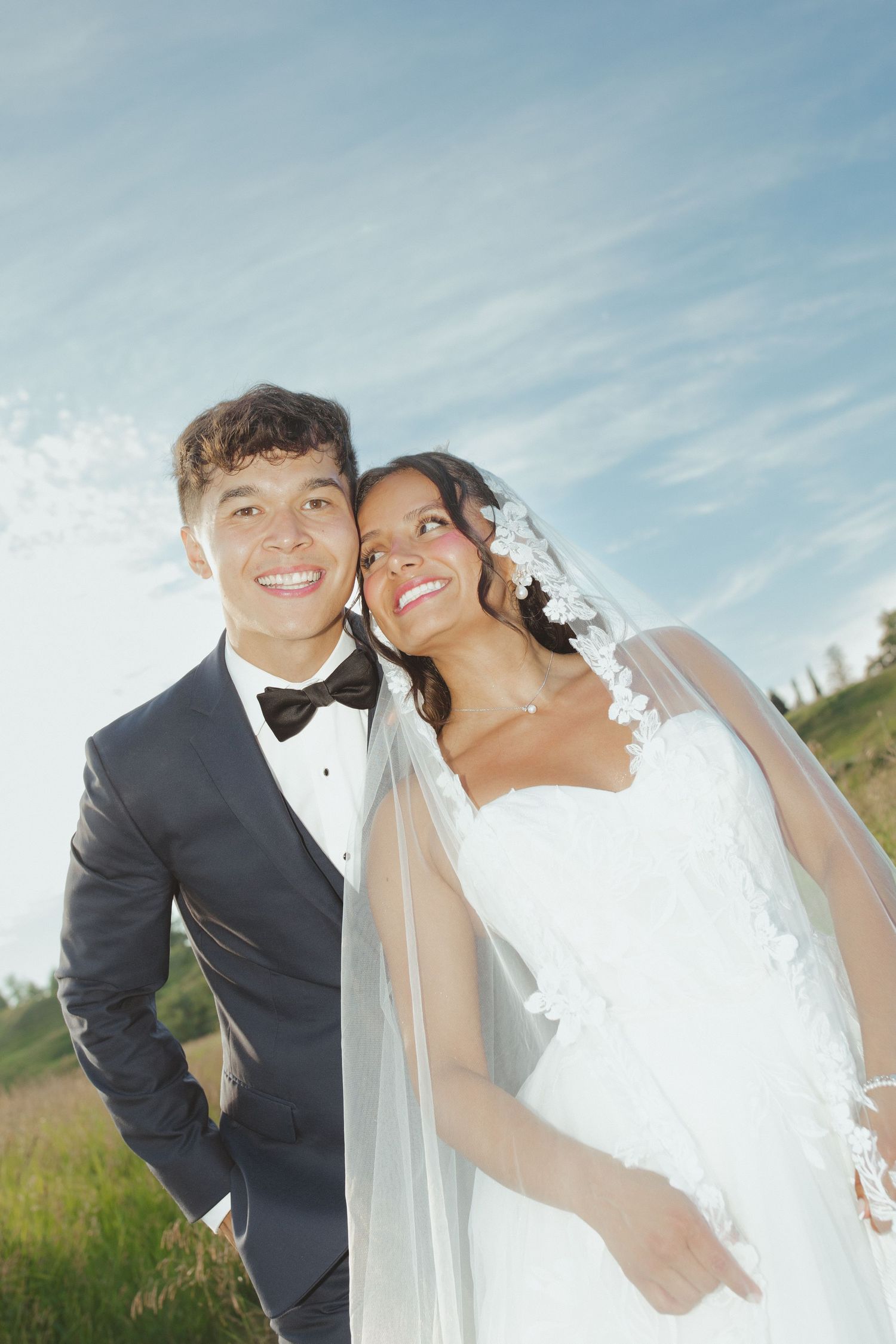 Smiling newlywed couple in formal attire, groom in tuxedo and bride in white gown with veil, posing outdoors under blue sky.