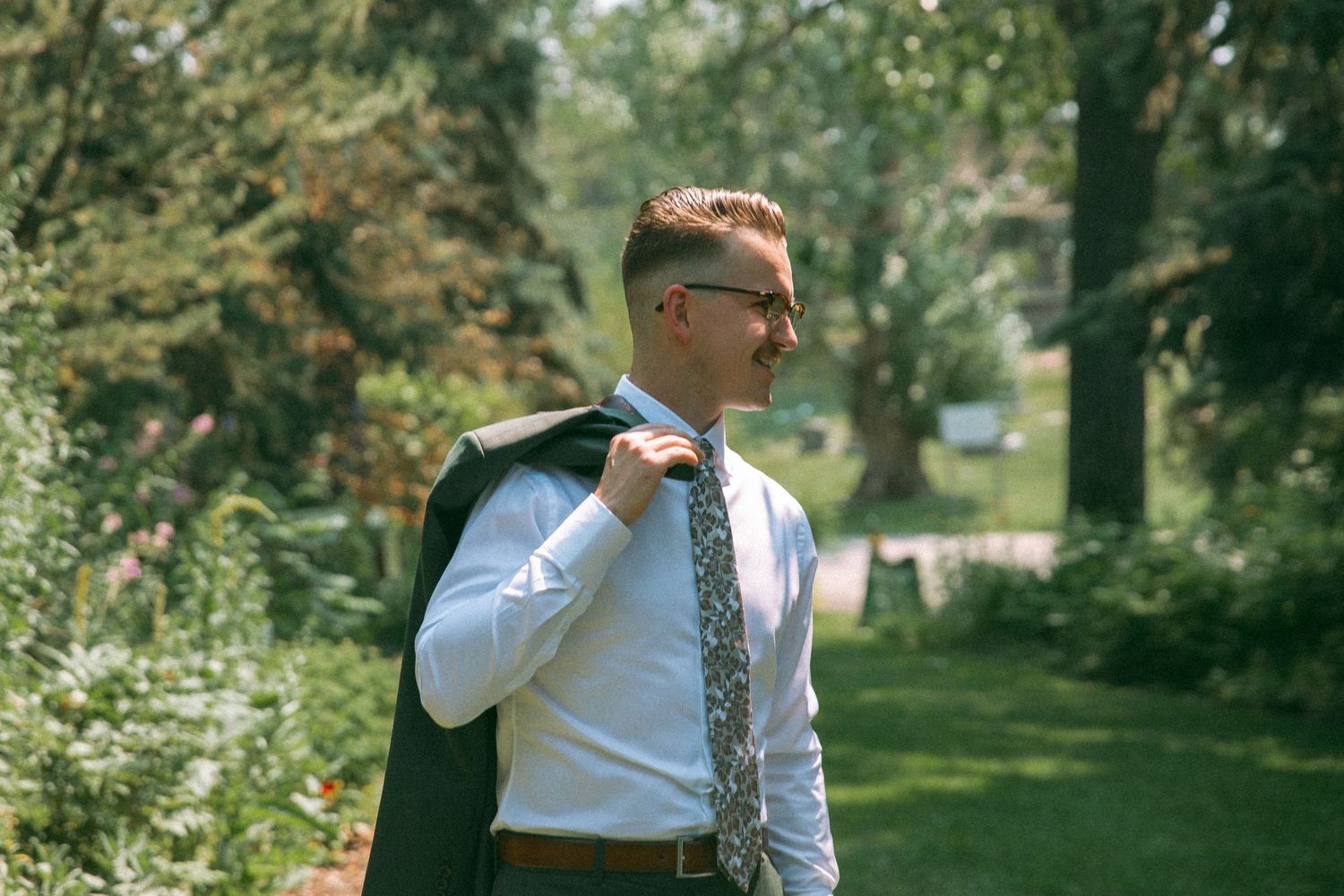 Groom in formal attire adjusts his jacket in a sunlit garden setting.