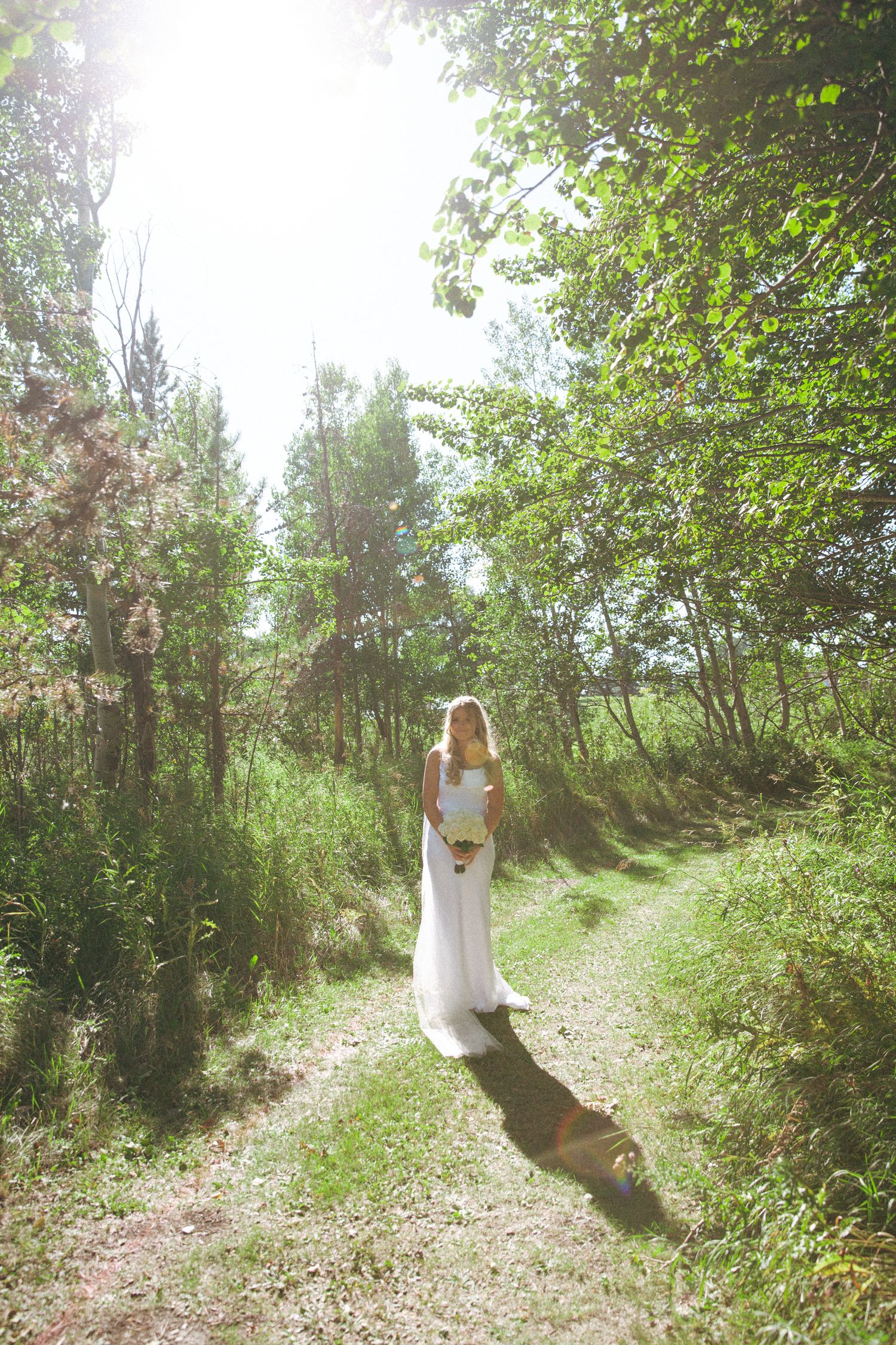 A bride in a white dress stands alone on a sunlit forest path holding a bouquet.
