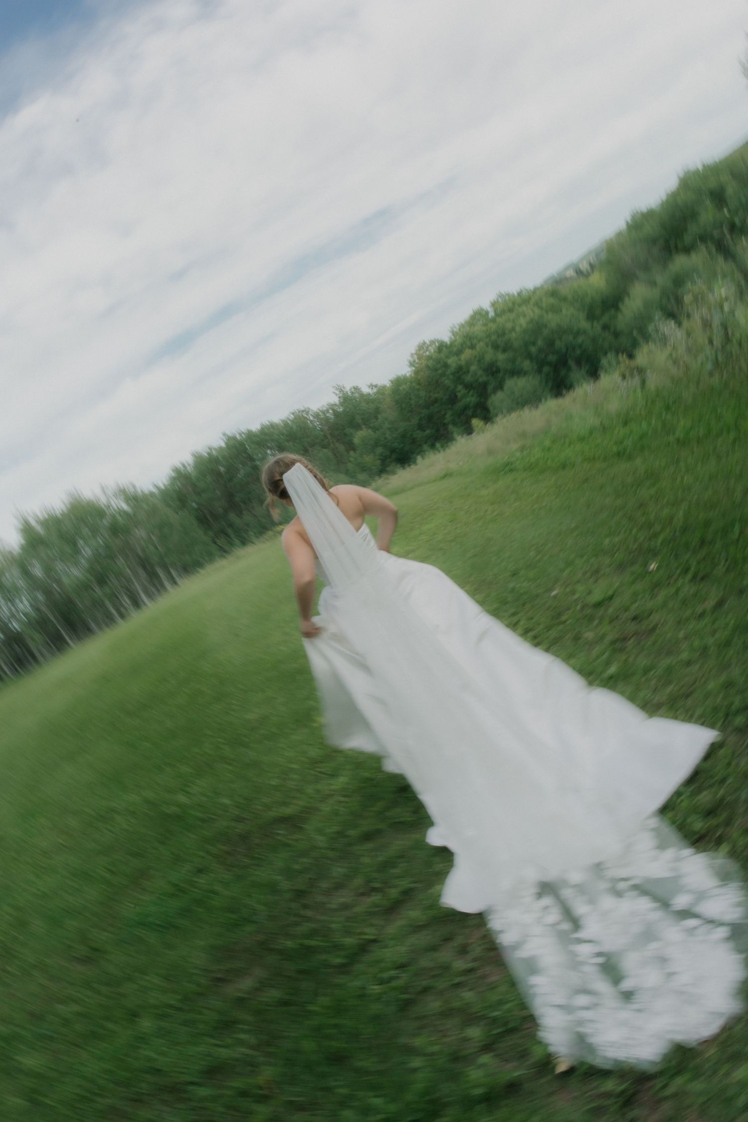 Bride in white gown and veil running through lush green field on wedding day.
