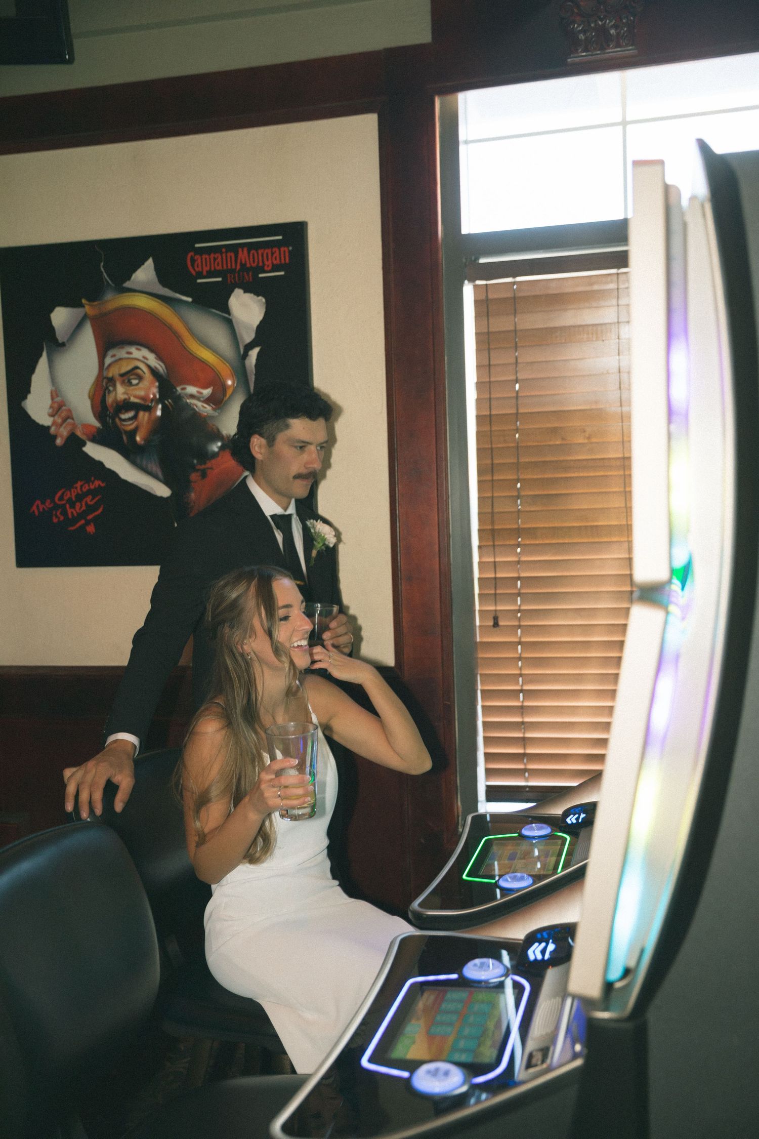 Bride and groom laughing at a slot machine in a bar, with a Captain Morgan poster on the wall behind them.
