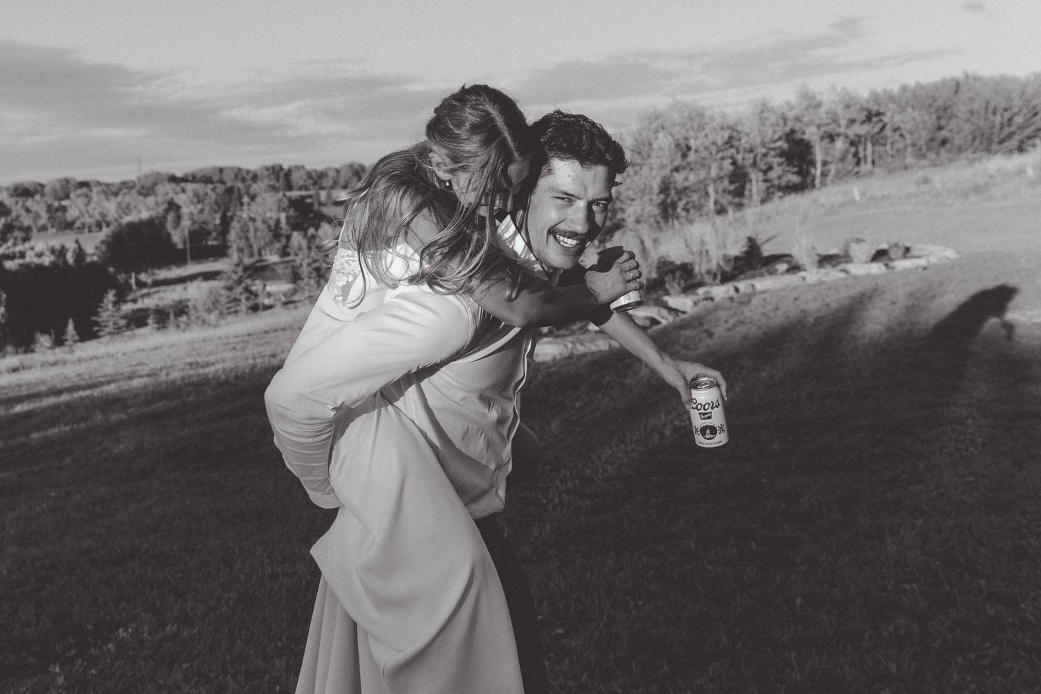 Black and white photo of a smiling man giving a woman a piggyback ride outdoors at a wedding celebration.