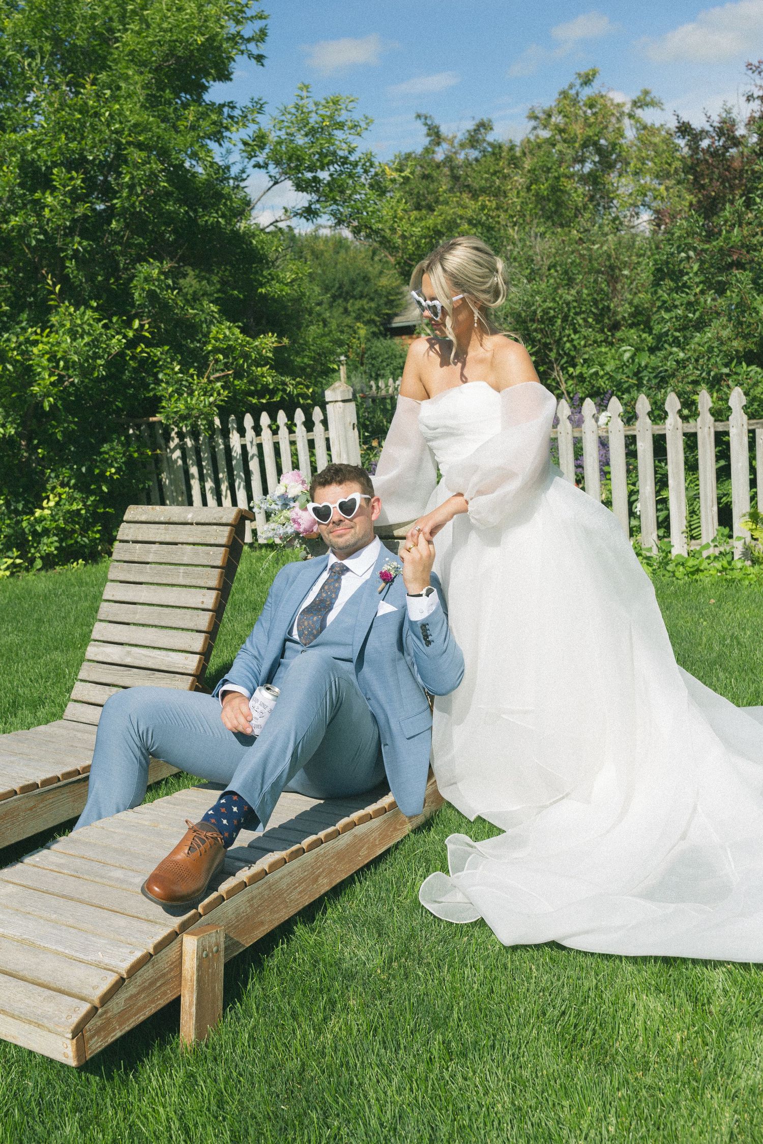 Wedding couple relaxes on wooden lounge chairs in a garden setting with white picket fence.