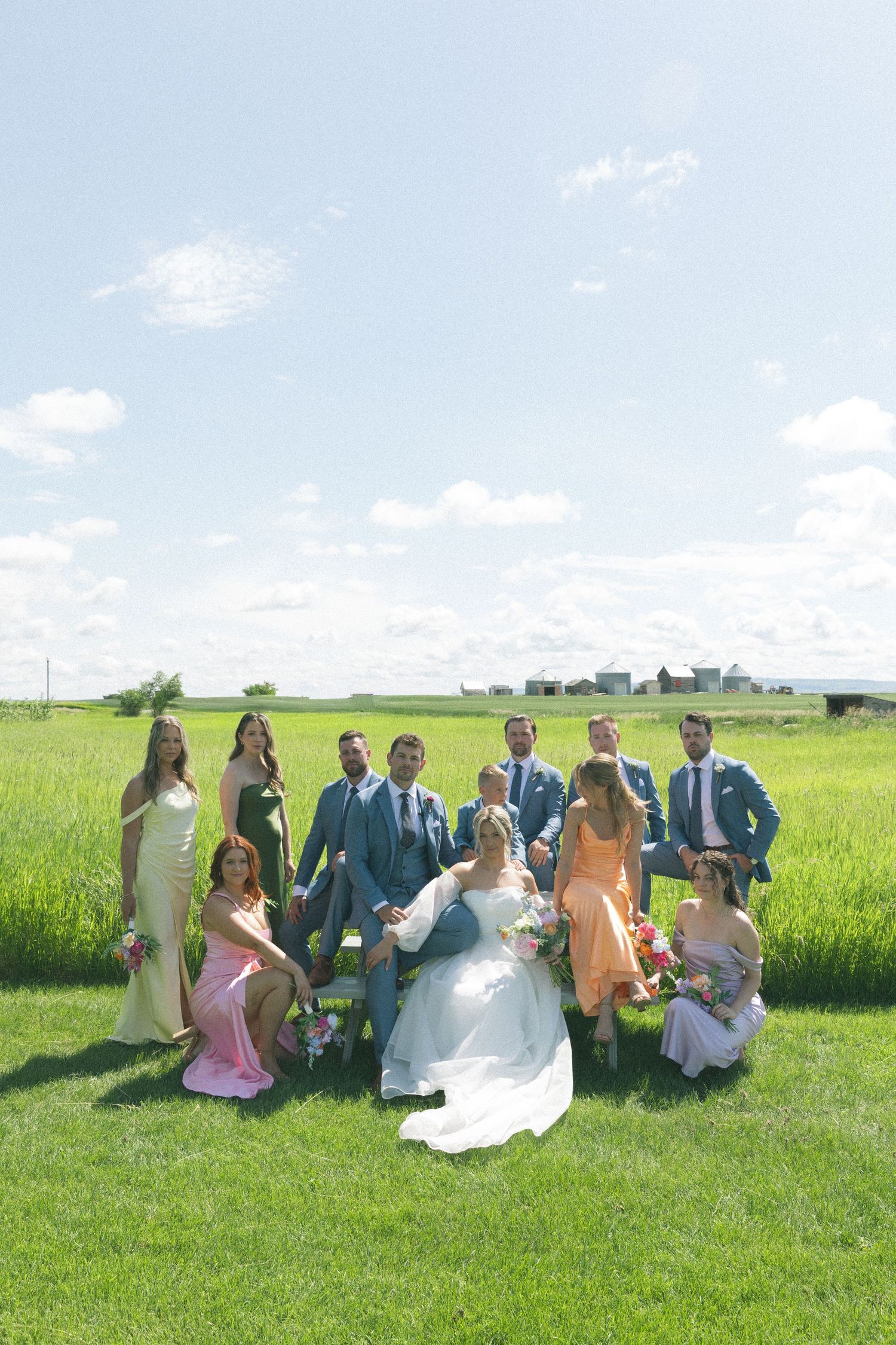 Wedding party poses for photos in a sunny green field with blue sky overhead.