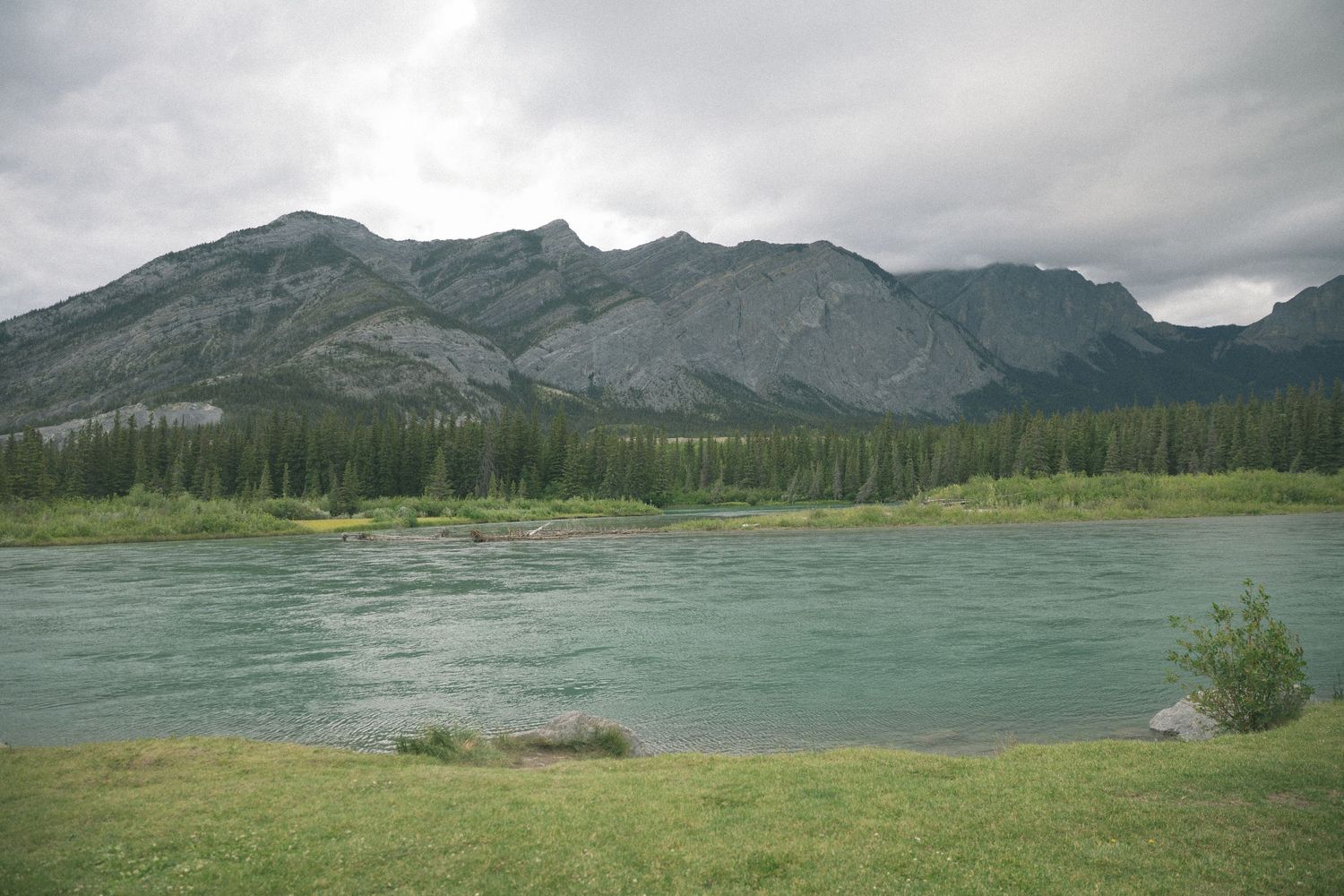 A scenic mountain landscape with turquoise river flowing through evergreen forest under cloudy skies.