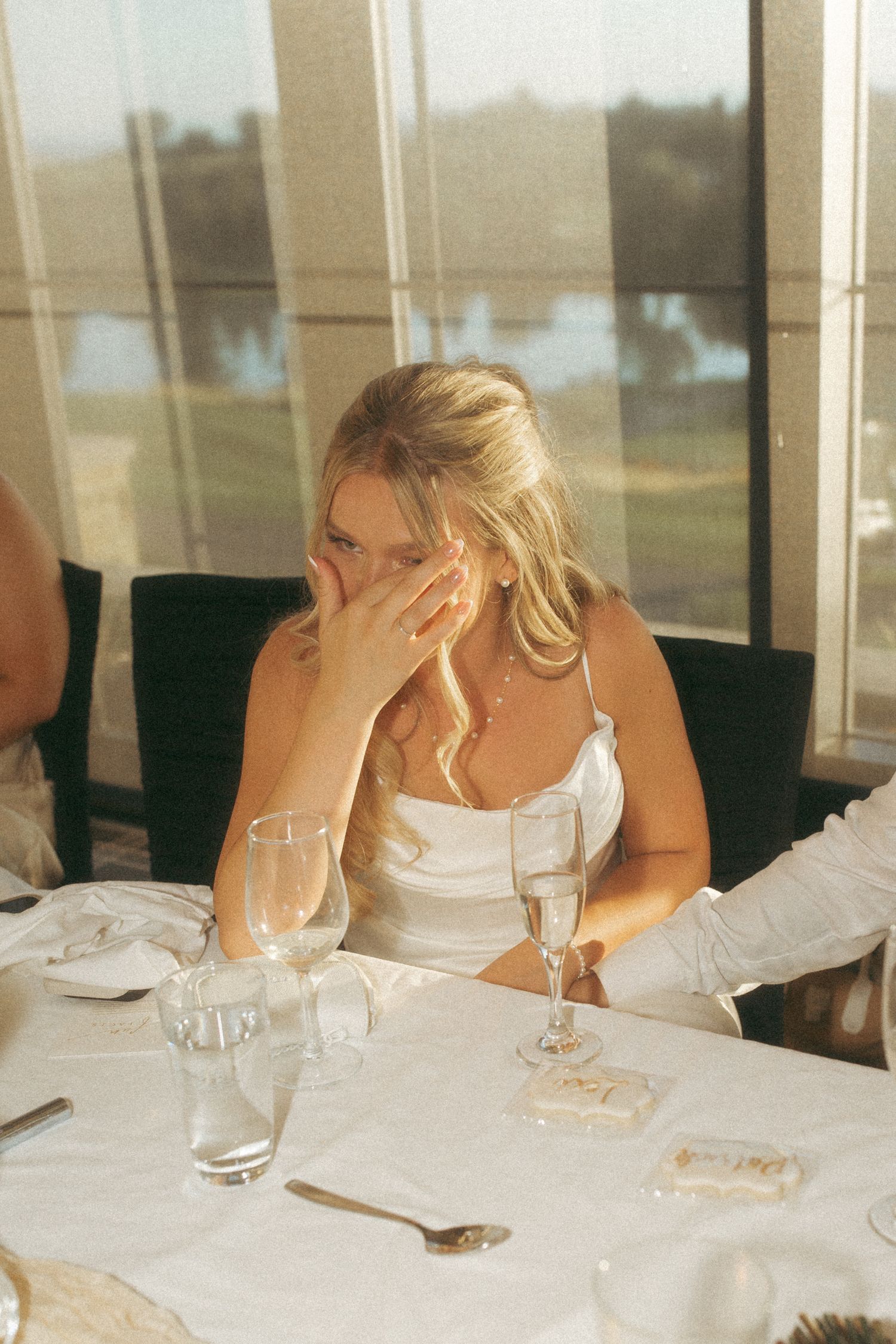 A person in a white dress rests their head on their hand at a formal dinner setting.