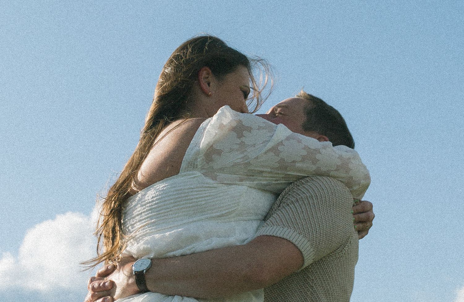 Couple in romantic embrace against blue sky as wind blows through long hair.
