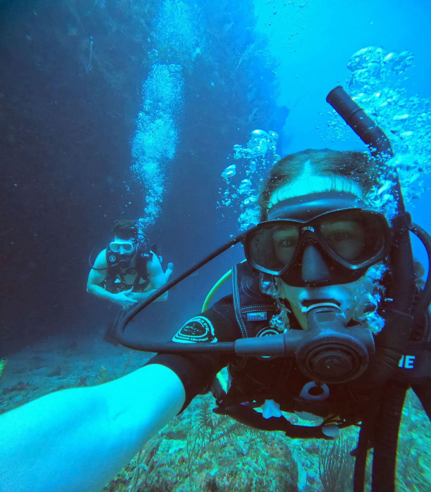Scuba diver takes underwater selfie in bright blue water with bubbles rising in background.