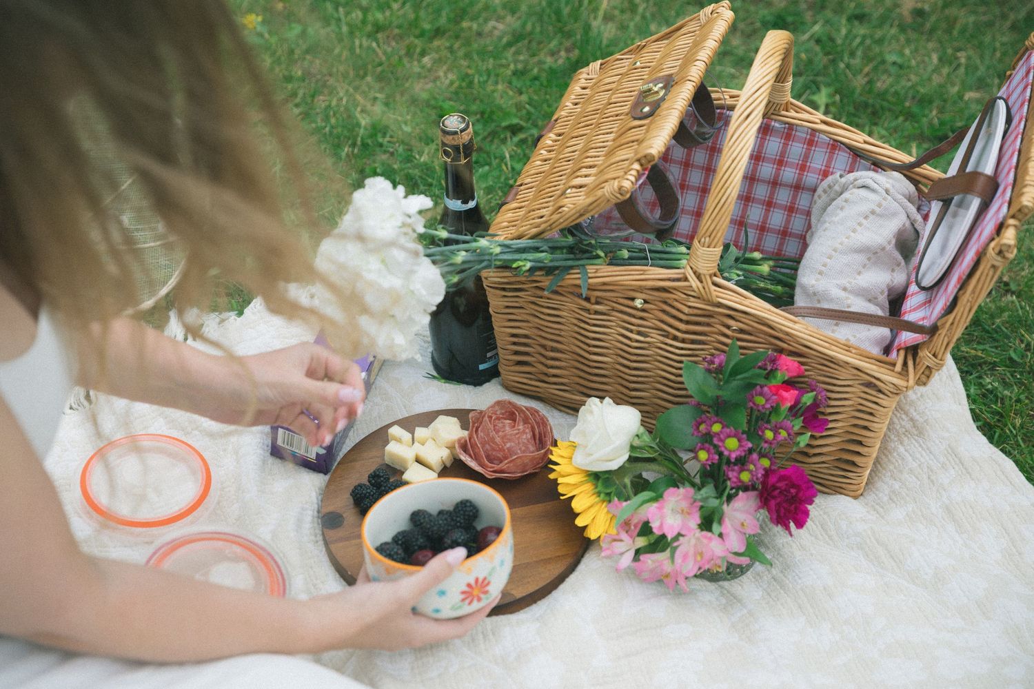 A summer picnic setup with a wicker basket, blanket, flowers, and a plate of fresh berries on the grass.