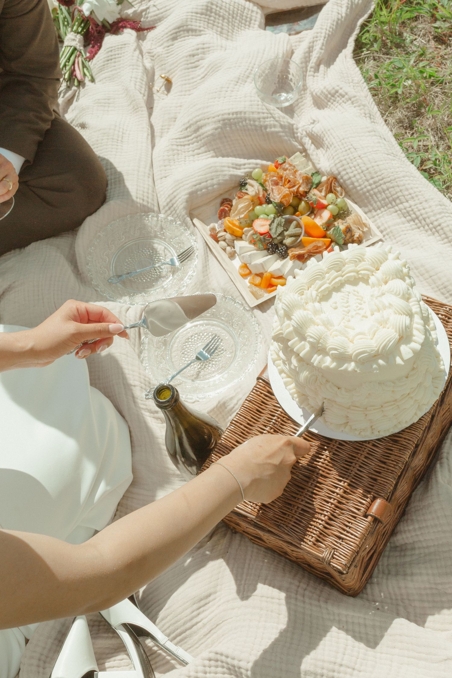 Woman cutting white layered cake at outdoor picnic with charcuterie board, wine bottle, and glasses on blanket.