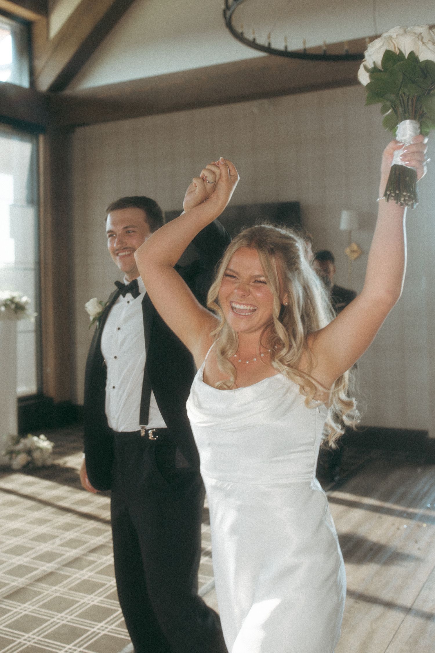 A bride in a white dress raises her arms joyfully while dancing at a wedding reception.