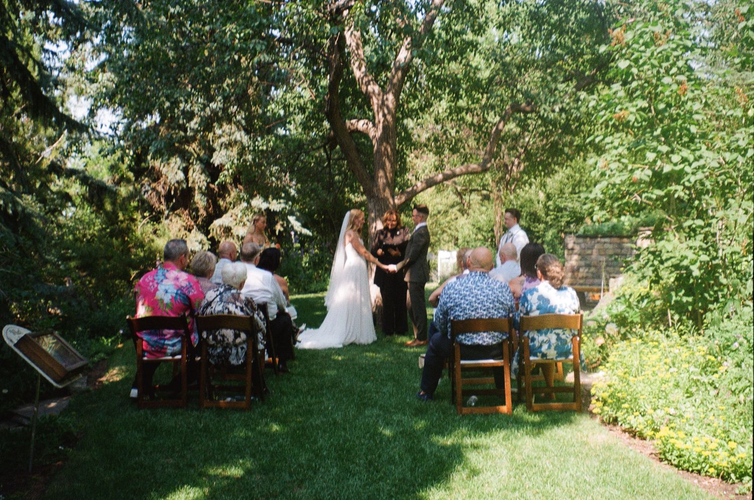 An intimate outdoor wedding ceremony takes place in a lush garden setting with wooden chairs arranged on a grassy lawn.
