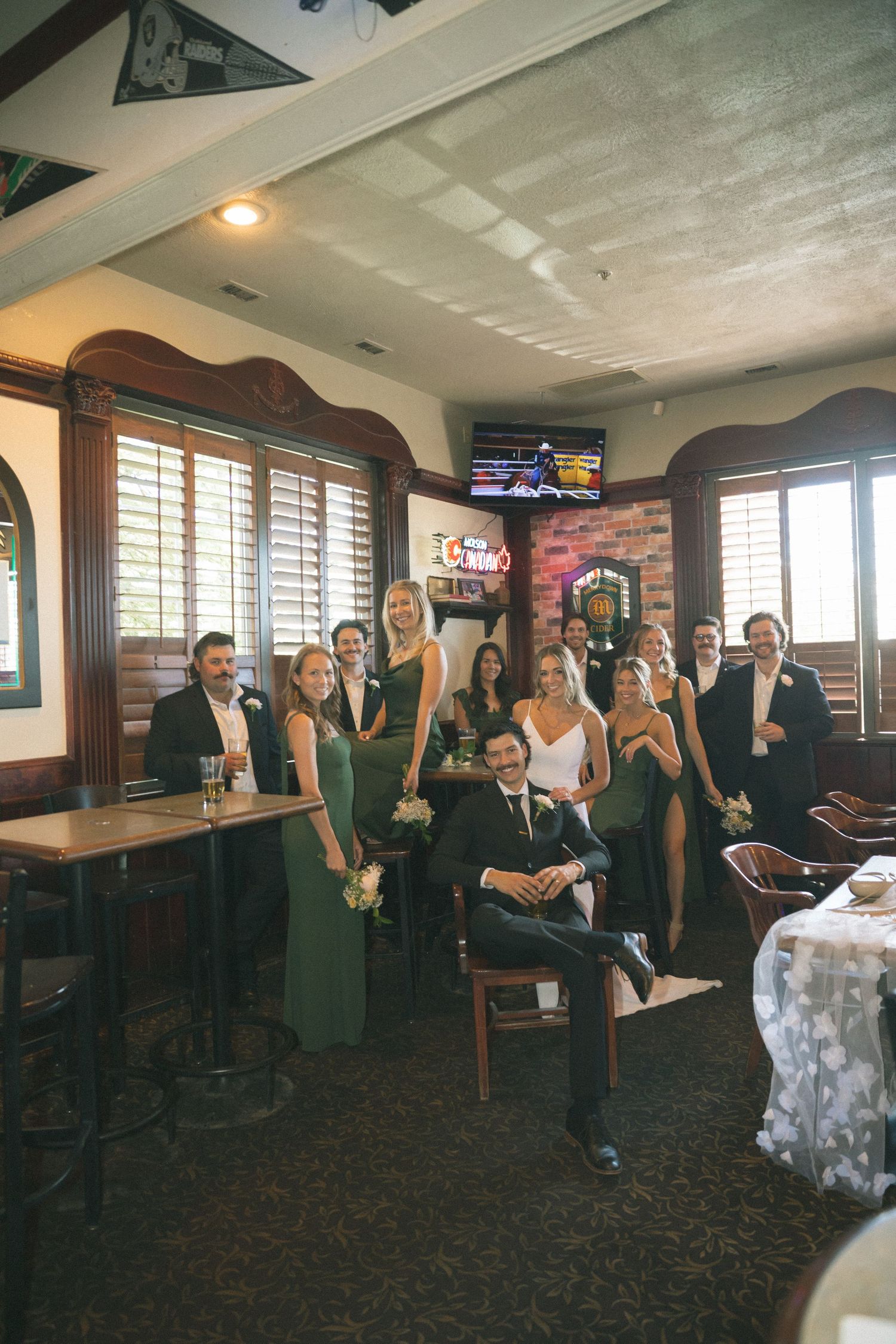 Wedding party poses together in a rustic bar venue, bridesmaids in green dresses with groom seated center.