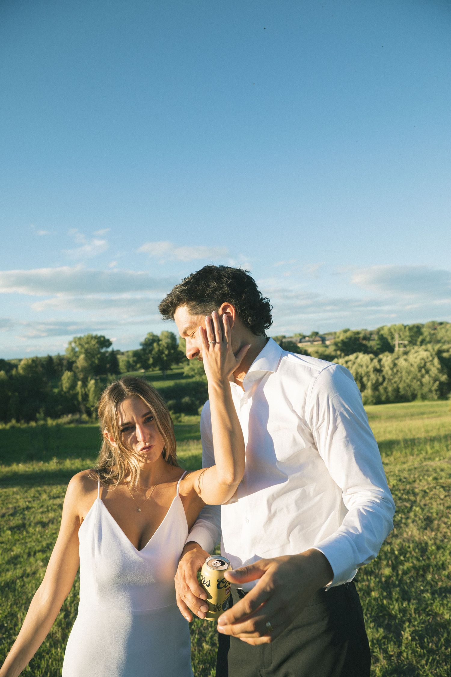 Bride and groom share a candid moment holding a beer can in a scenic green outdoor setting.