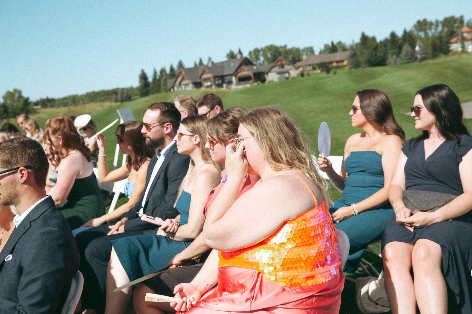 Wedding guests seated outdoors on white chairs for ceremony with scenic golf course backdrop.