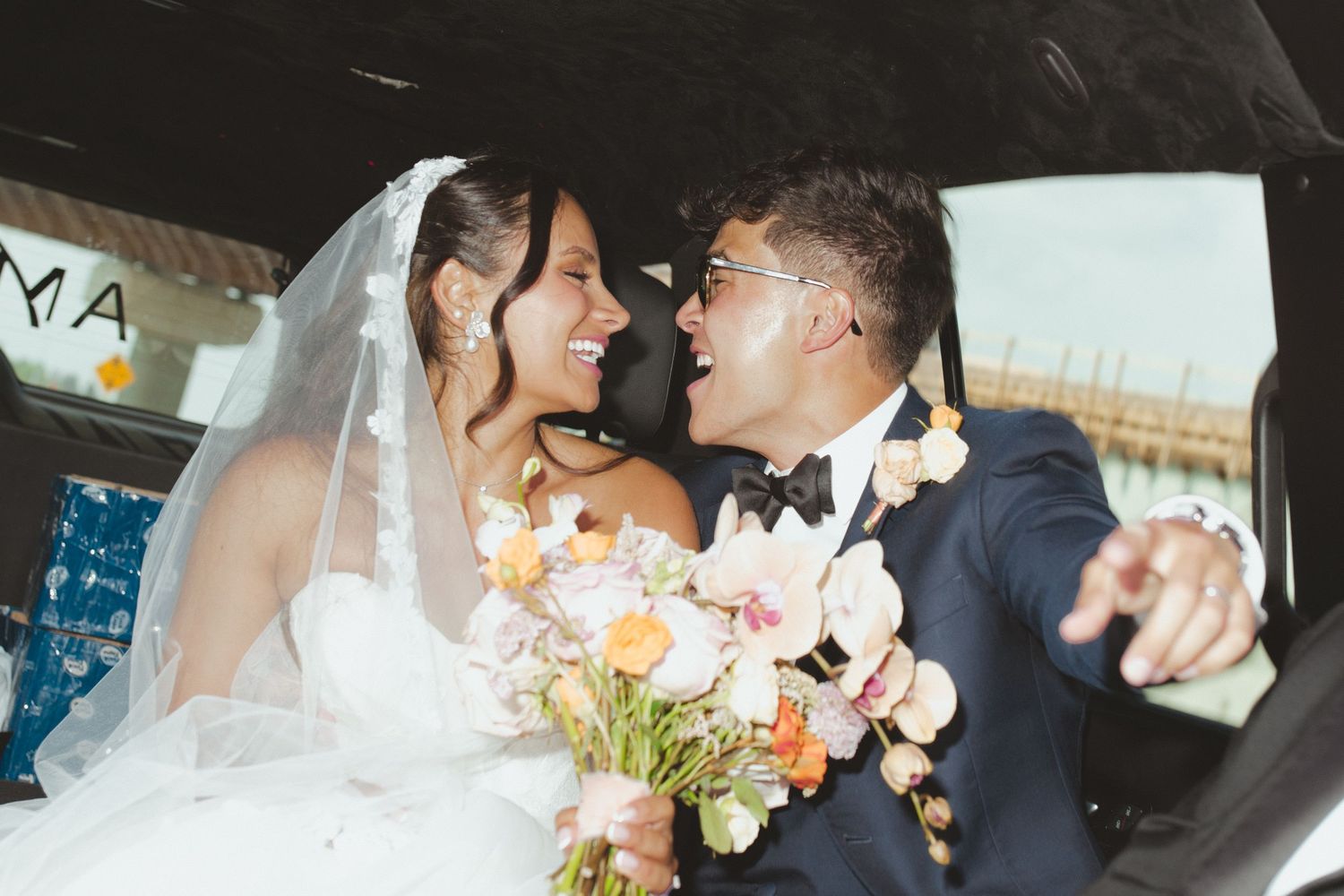 Bride and groom laughing together inside a car, bride holding a colorful bouquet on their wedding day.