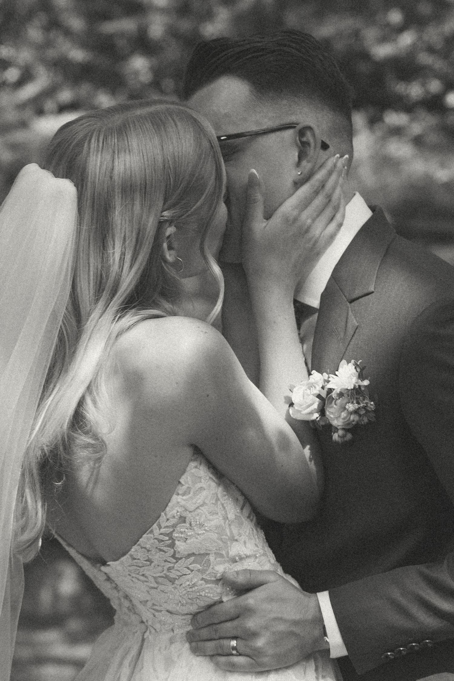 Intimate black and white wedding photo of bride in lace dress and groom in suit sharing a tender moment outdoors.
