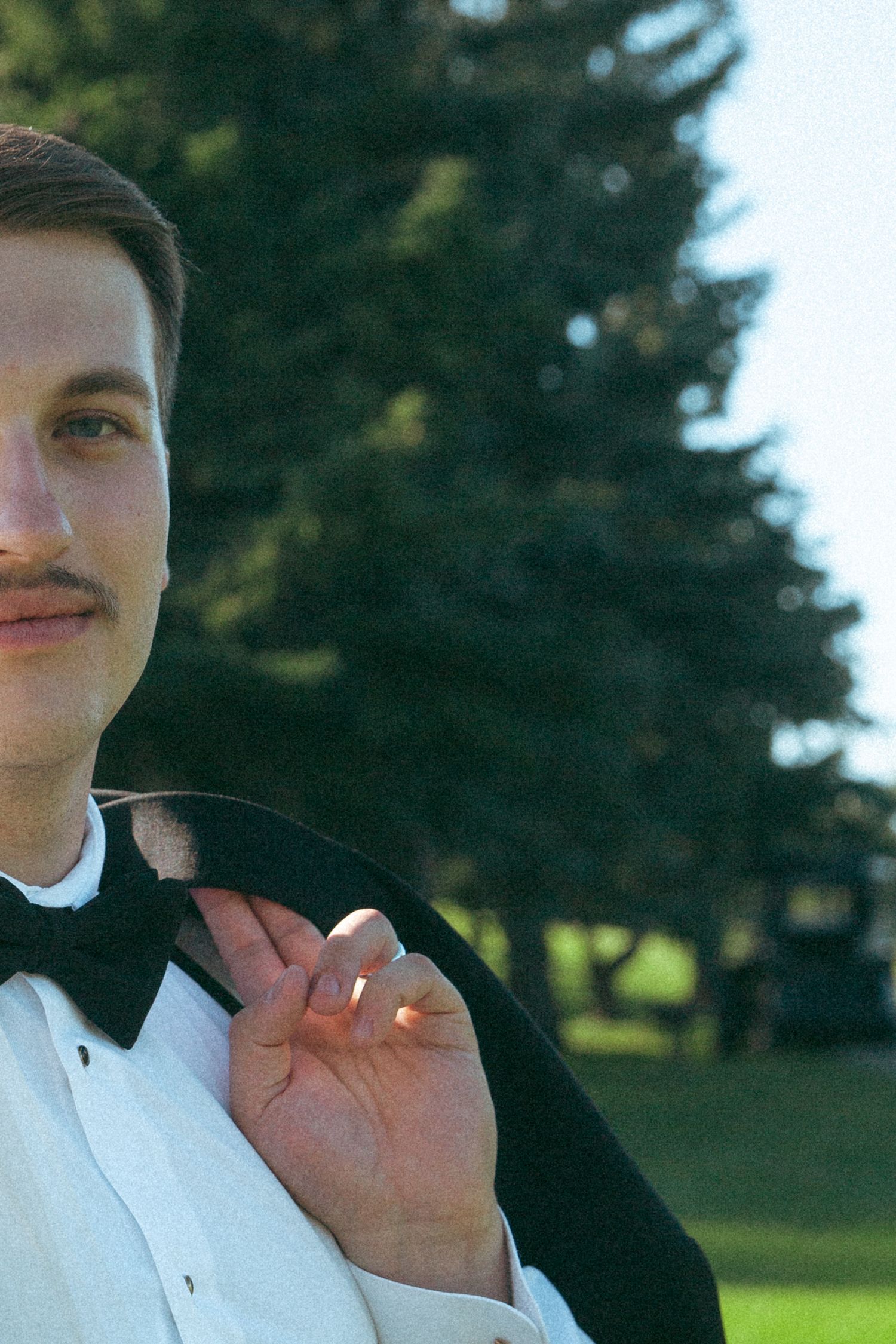 A groom in classic black tuxedo and bowtie poses against an evergreen backdrop.
