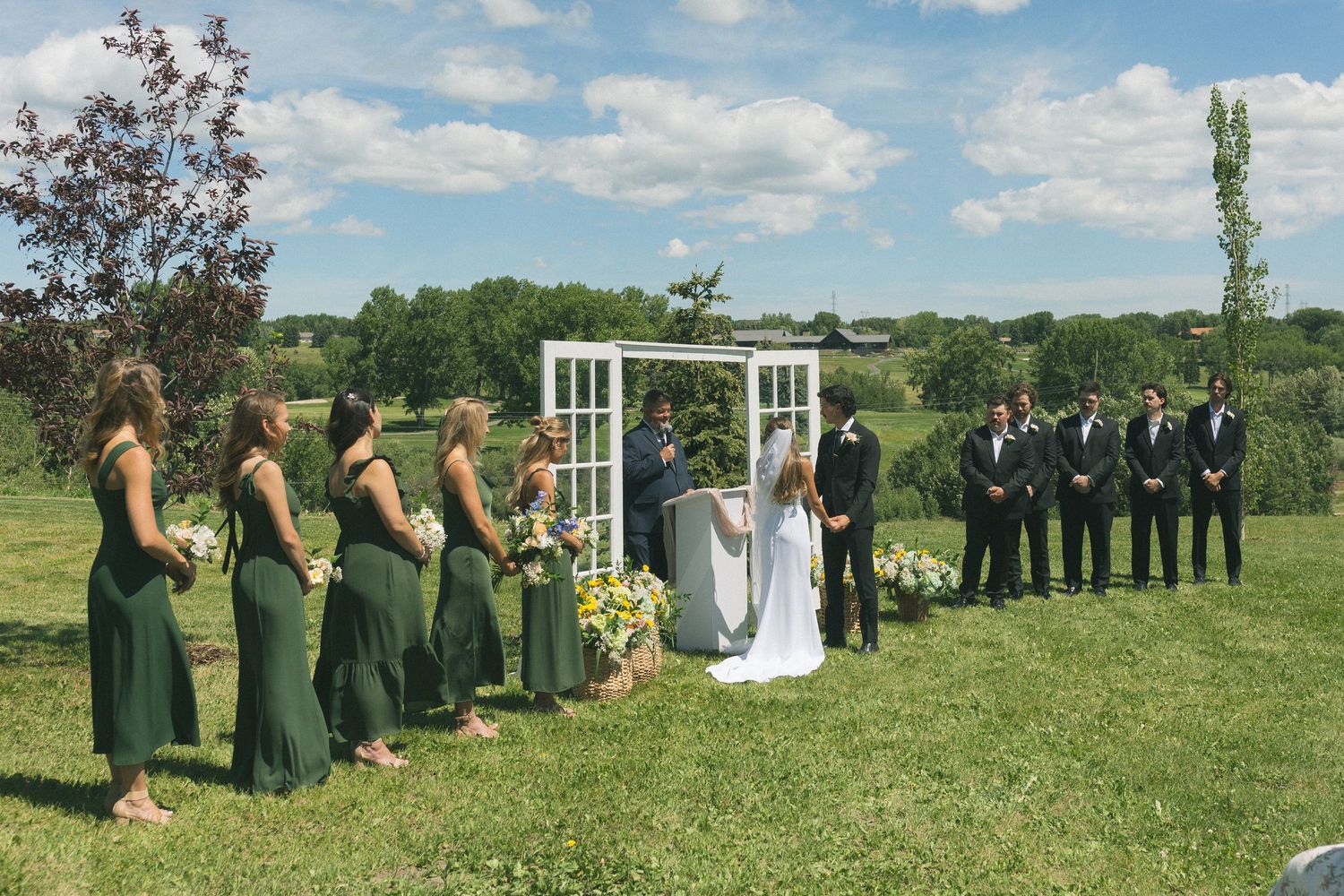 Outdoor wedding ceremony on green lawn with bridesmaids in emerald dresses and groomsmen under a white door arch.