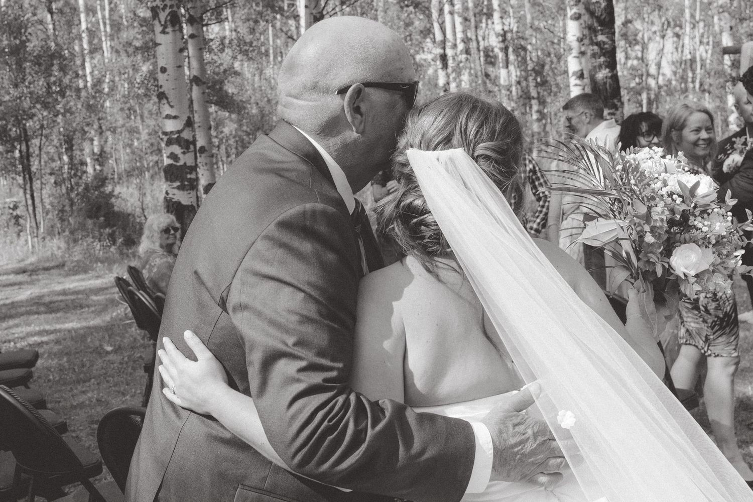 Black and white photo of a bride and groom sharing their first kiss at an outdoor wedding ceremony among birch trees.