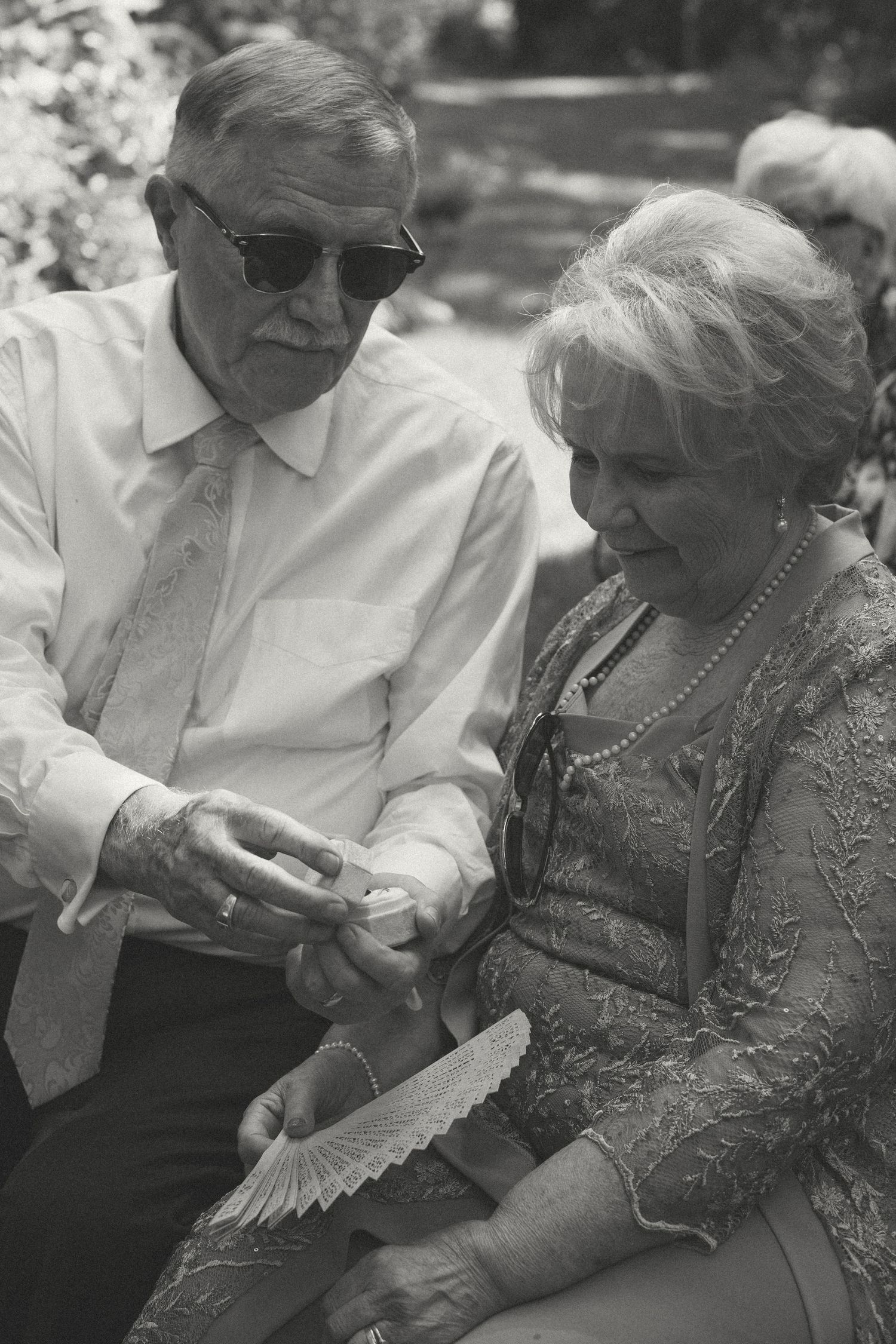 An elderly couple sits together at outdoor wedding giving blessing to wedding rings.