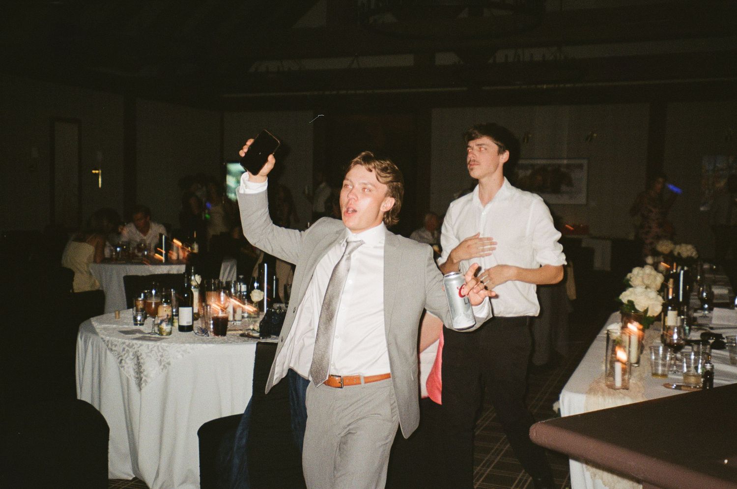 Man in grey suit and tie celebrating enthusiastically at a reception with round tables and other guests in background.