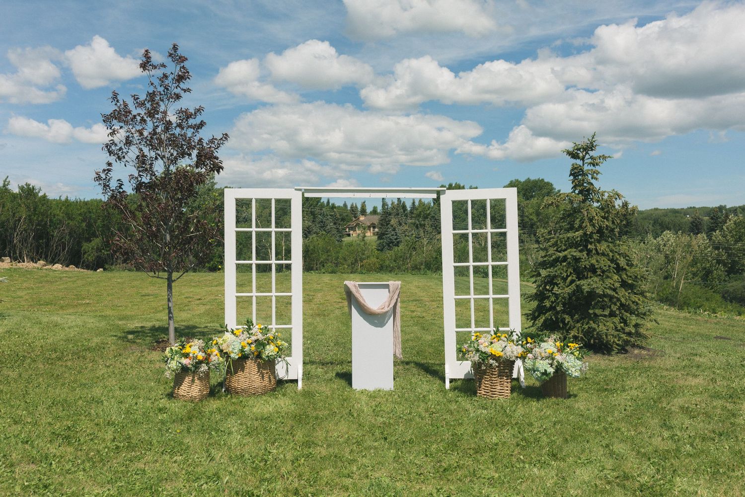Rustic outdoor wedding altar with white French doors, floral arrangements, and a pedestal on a sunny green lawn.