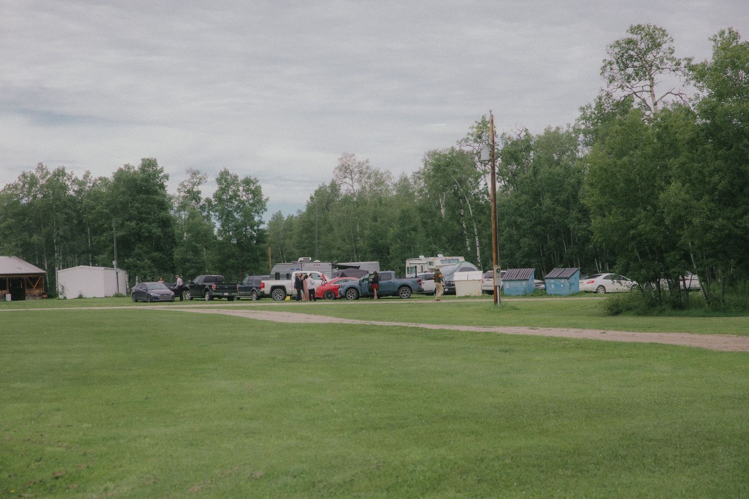 Rustic campground with RVs and trucks parked among green trees on a cloudy day.