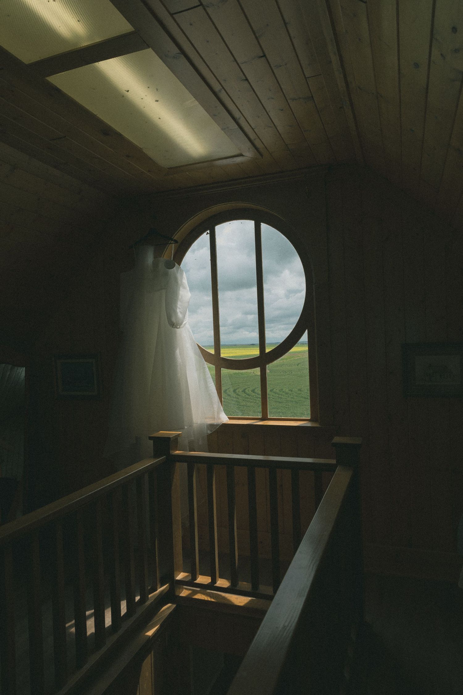 Round window in dark wooden stairwell reveals scenic countryside view with white curtain.
