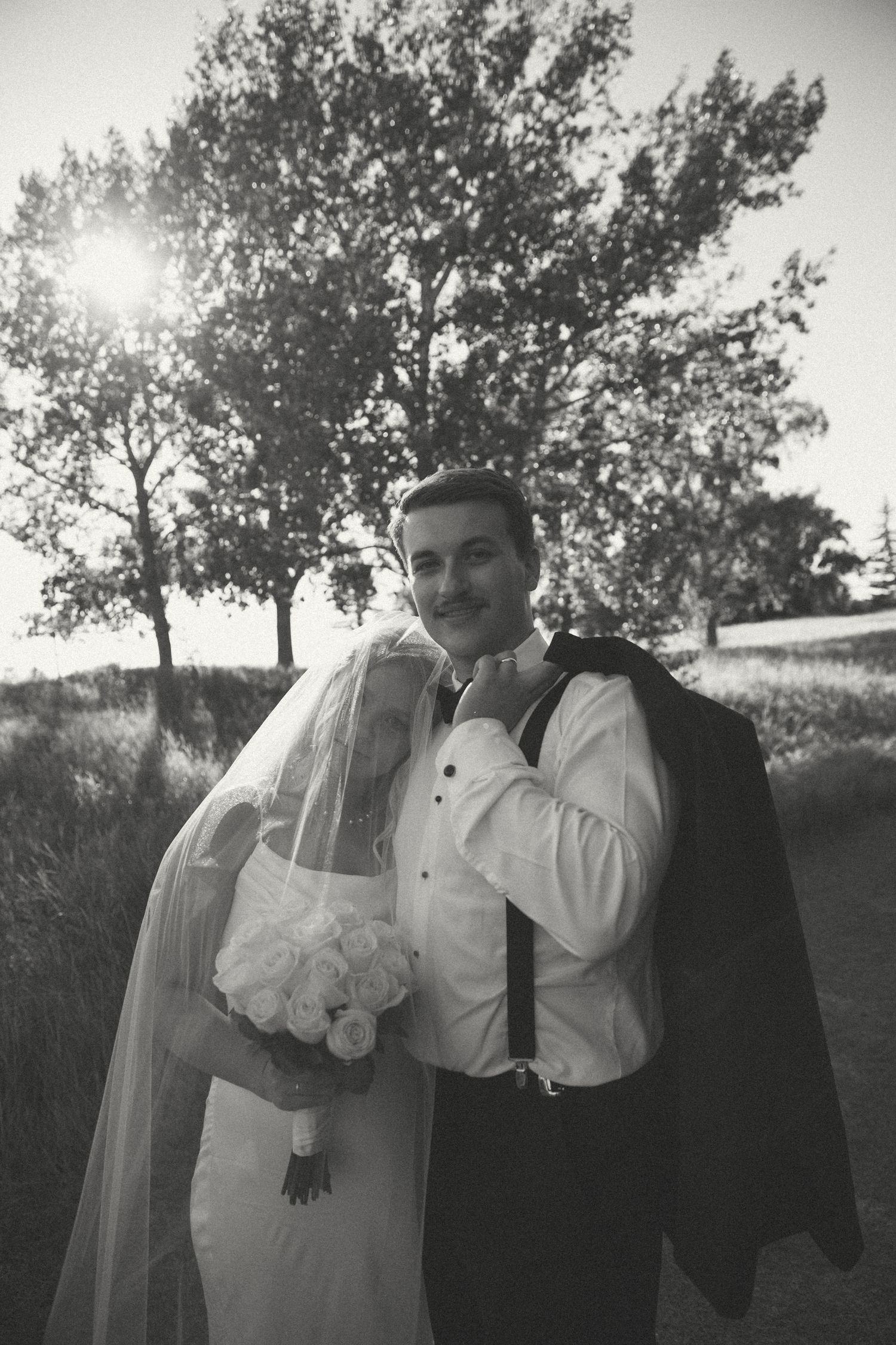 A black and white wedding portrait with dramatic sunlight filtering through trees.