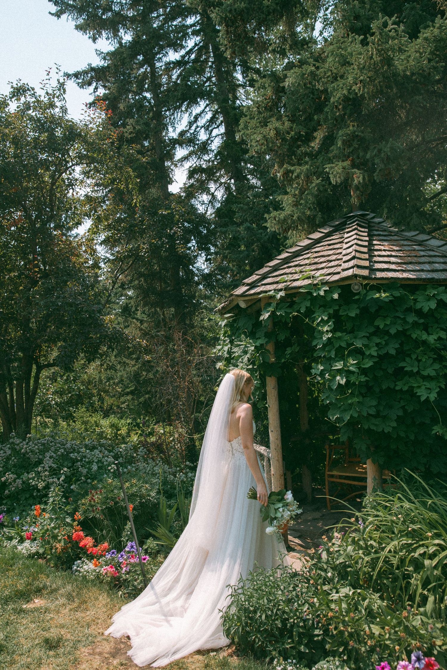 Bride in flowing veil stands by rustic garden gazebo surrounded by summer flowers.