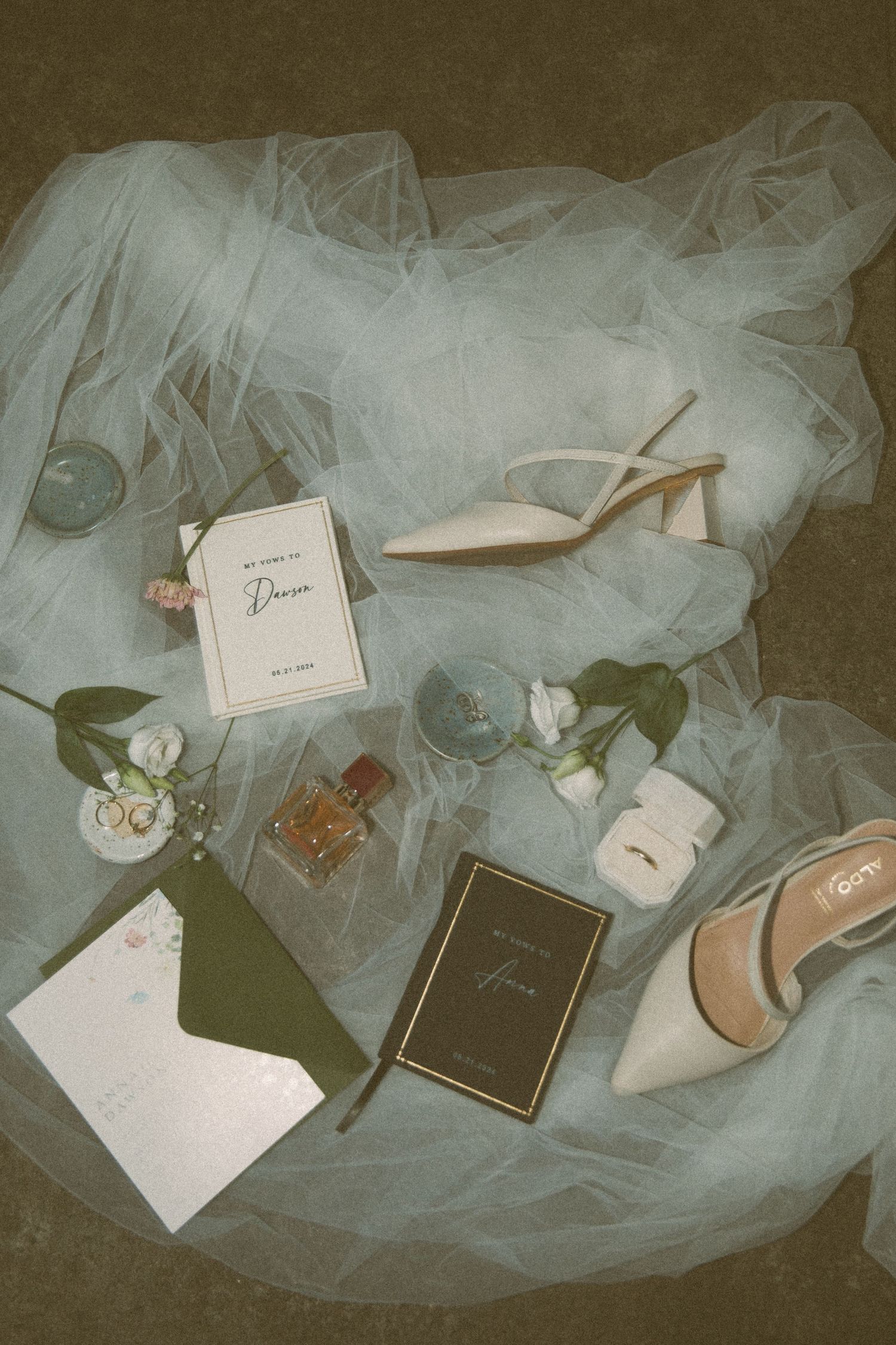 Elegant wedding accessories including white heels, perfume bottles, rings, and delicate tulle veil on a dark background.