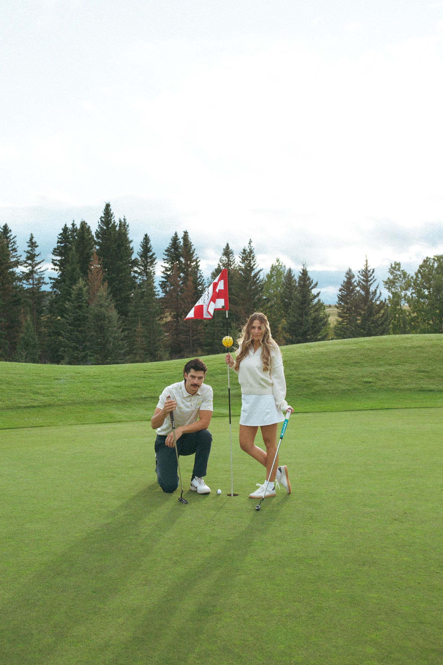 Couple poses by a golf flag on a beautiful course with Canadian flag and mountain backdrop.