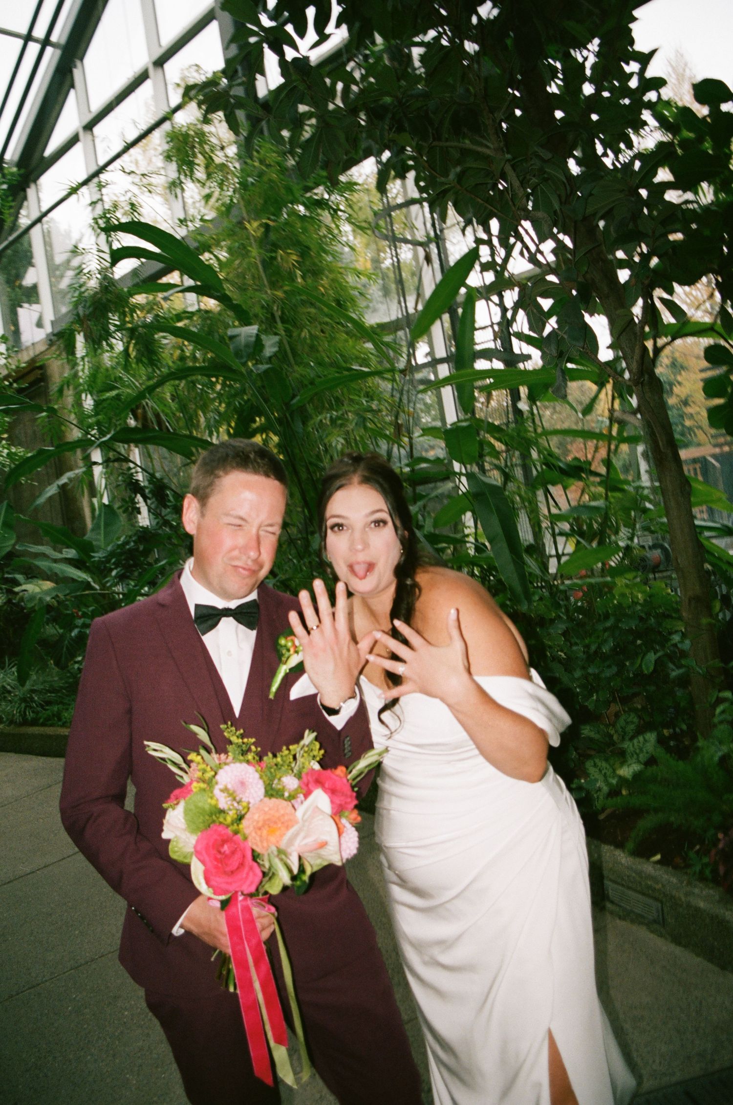 Newlyweds showing off wedding rings in a greenhouse, groom in burgundy tuxedo, bride in white gown holding bouquet.