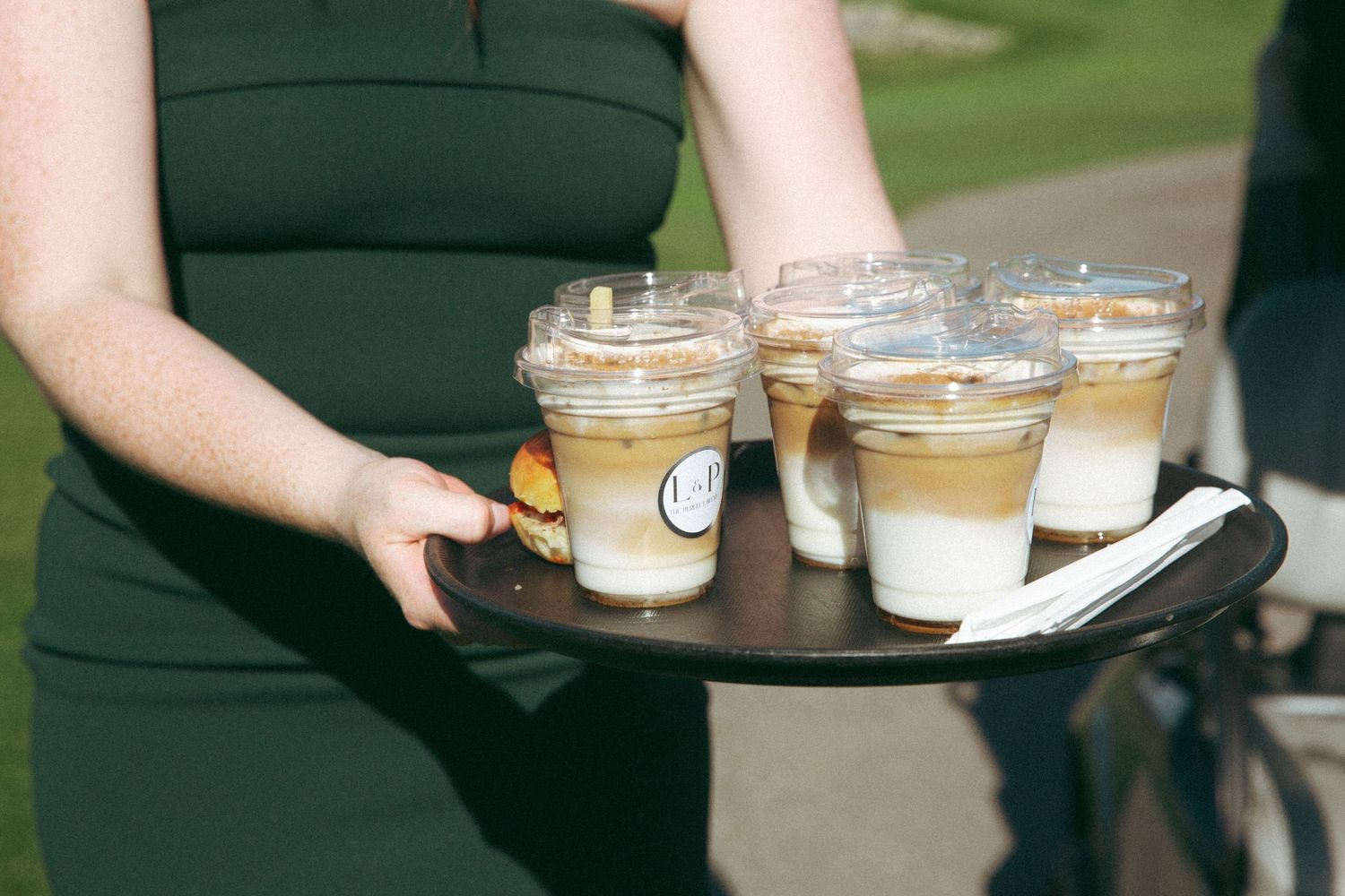 Server carrying tray of layered coffee drinks at outdoor event.
