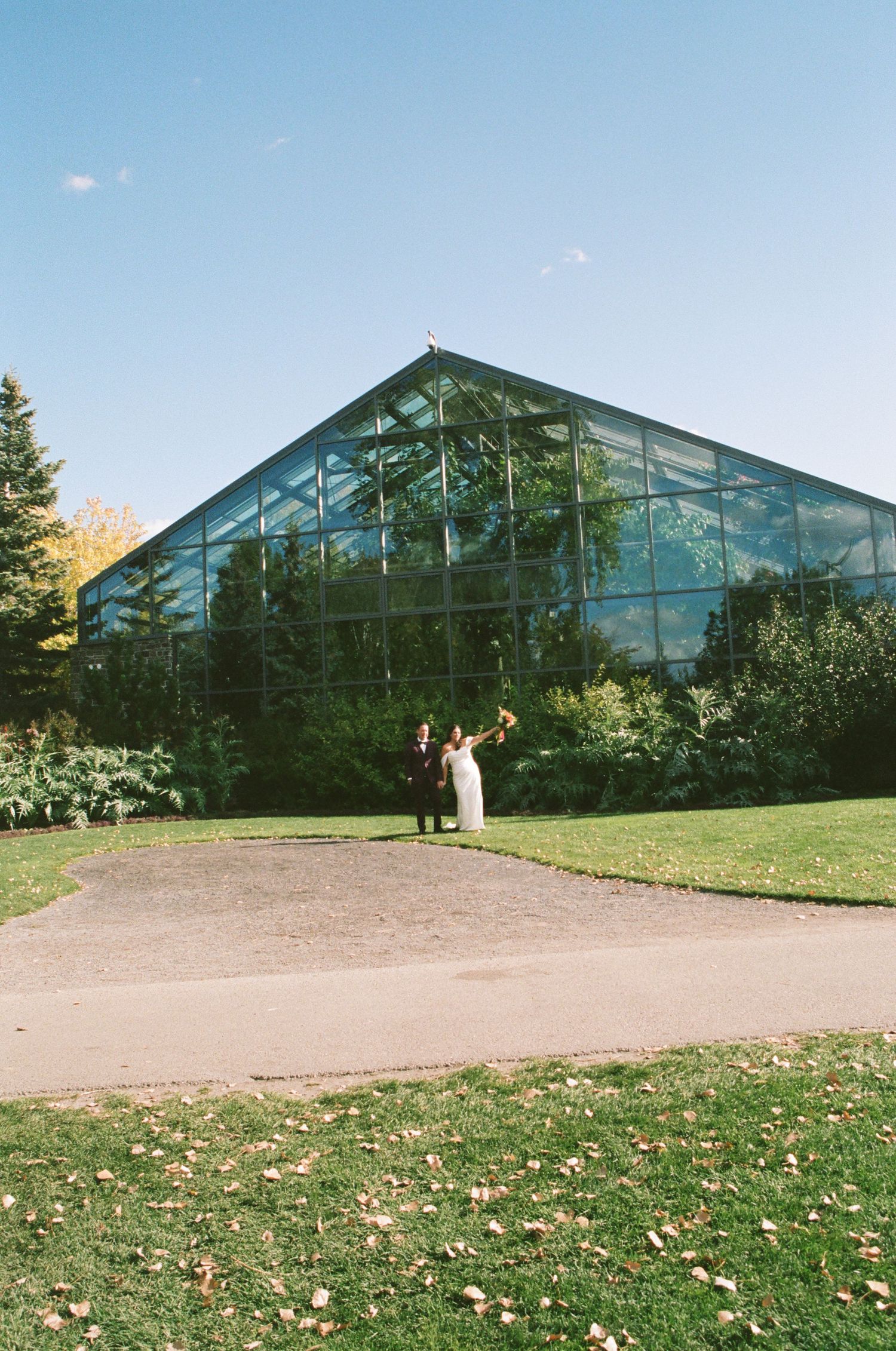 Wedding couple celebrating in front of a large glass greenhouse surrounded by green trees under a clear blue sky.