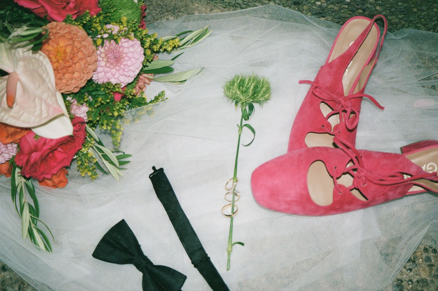 Pink suede bridal heels beside a colorful wedding bouquet, black bow tie, and rings on white tulle fabric.