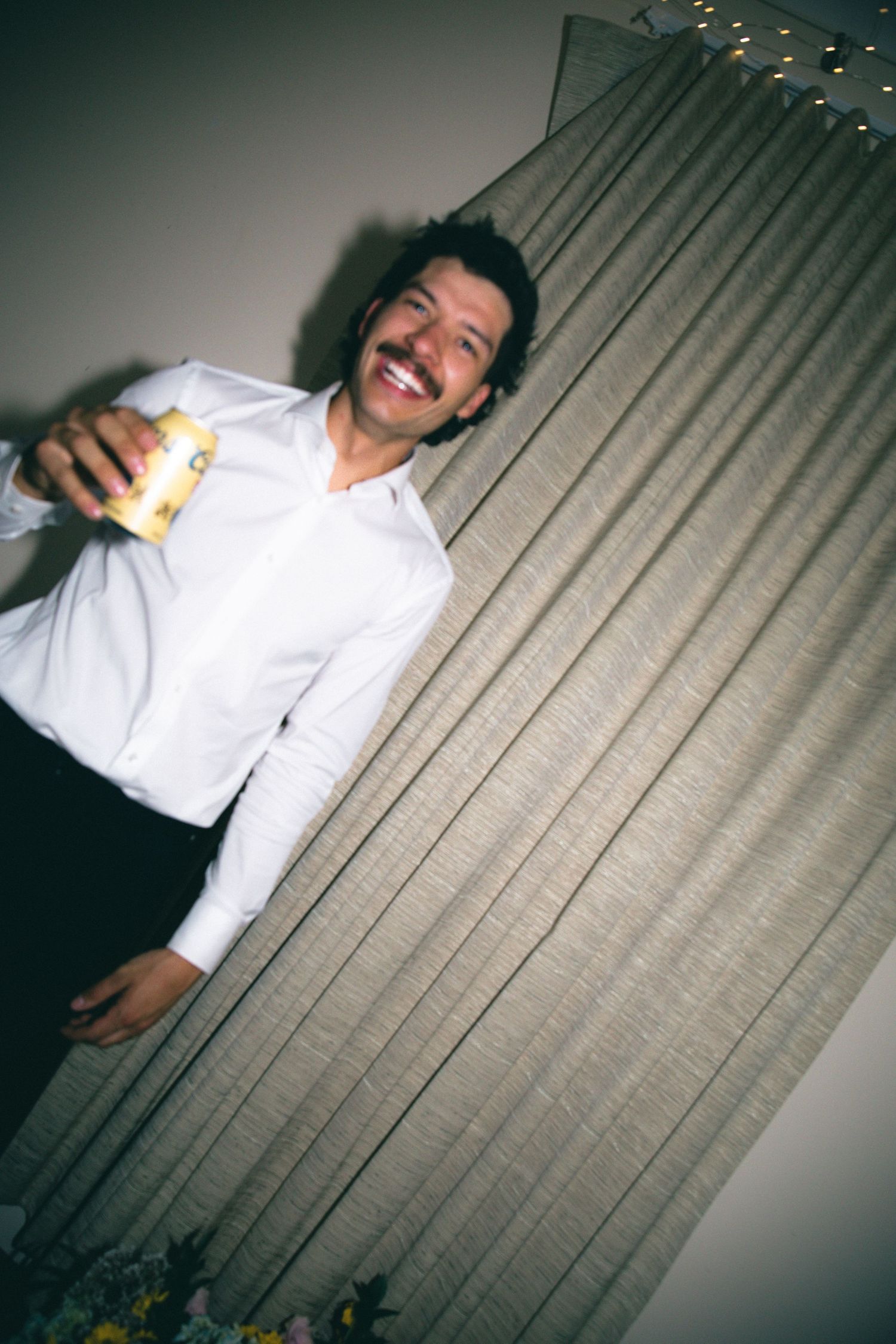 Smiling man with mustache in white shirt holding a beer can, standing by a gray curtain at a party.