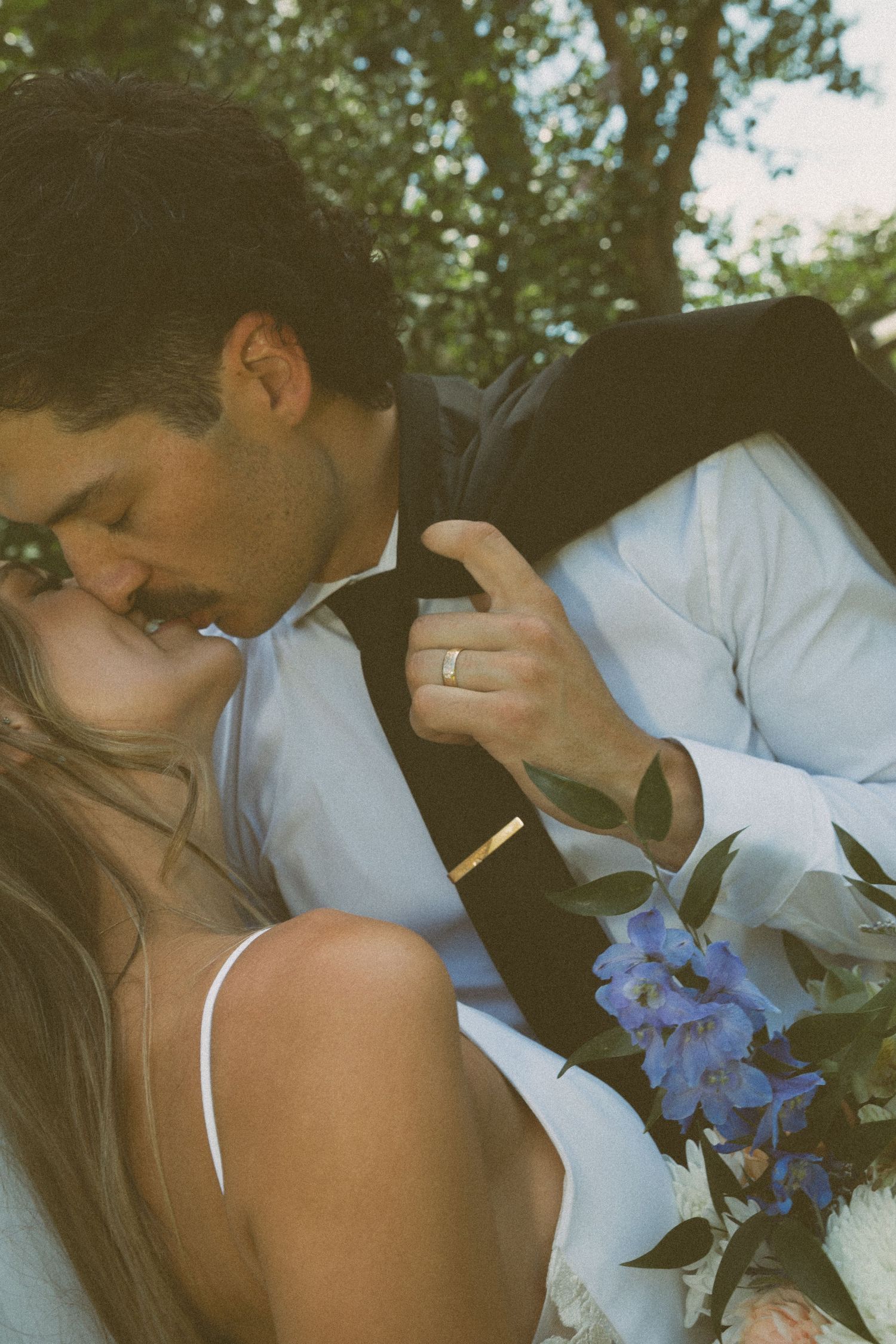 A bride and groom share a romantic kiss outdoors, with blue flowers and lush greenery in the background.