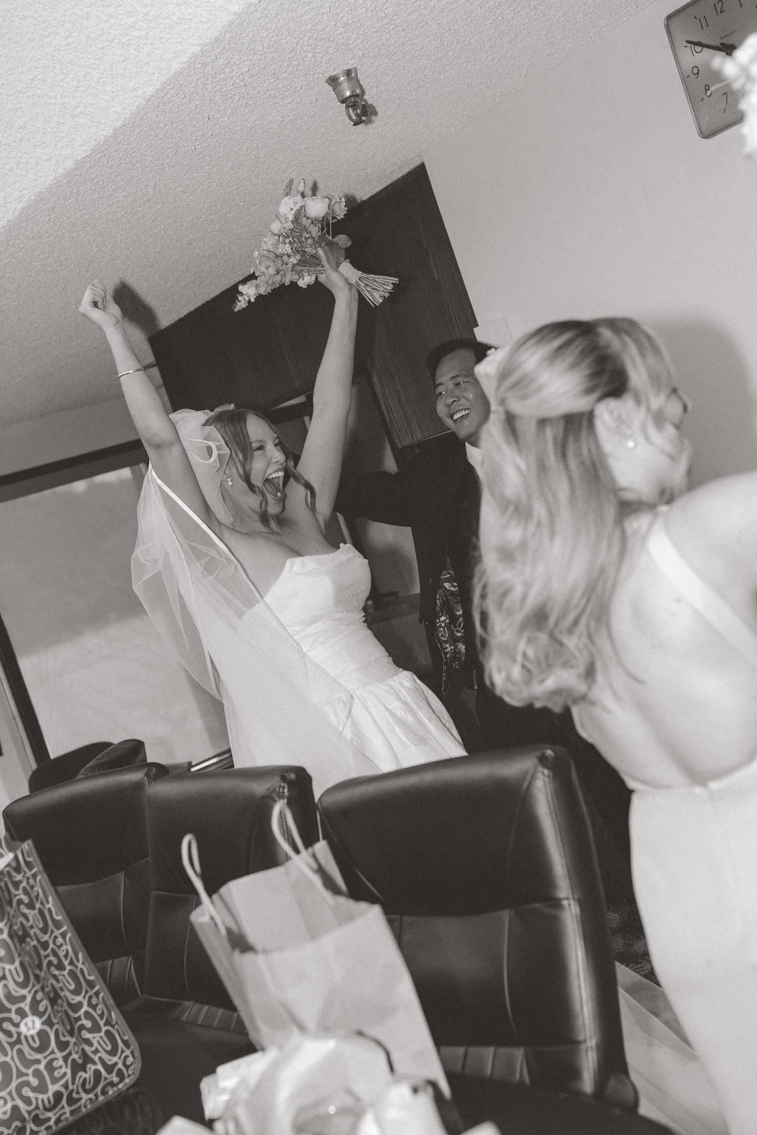 Bride in white gown raises bouquet joyfully with bridesmaids in black and white wedding photo.