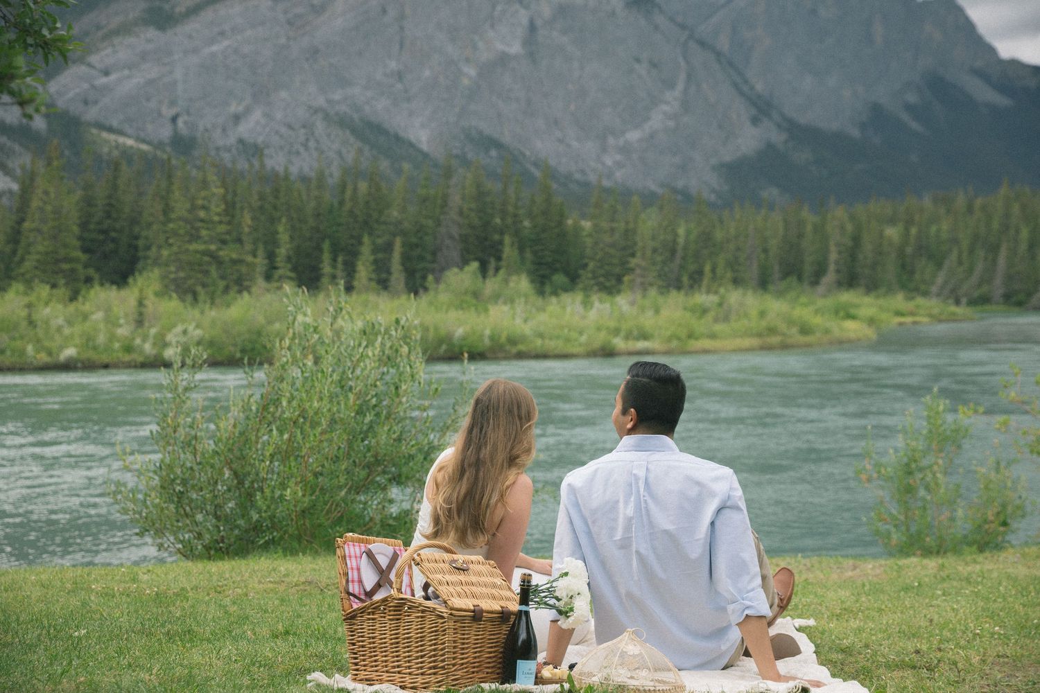 A couple enjoys a romantic picnic by a turquoise river with mountains and evergreen forest in the background.
