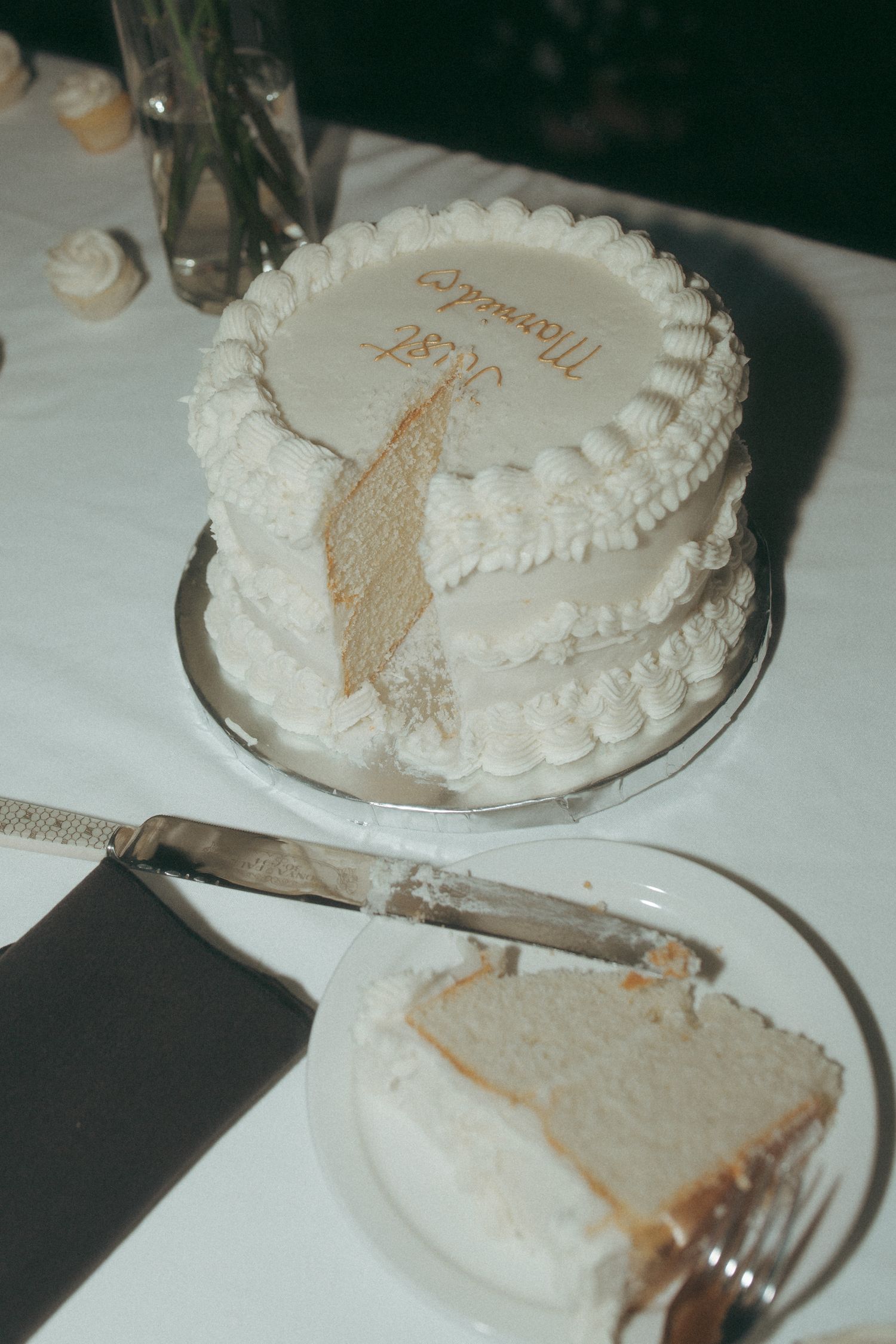 A white wedding cake with delicate piping sits on a table with a slice being served.
