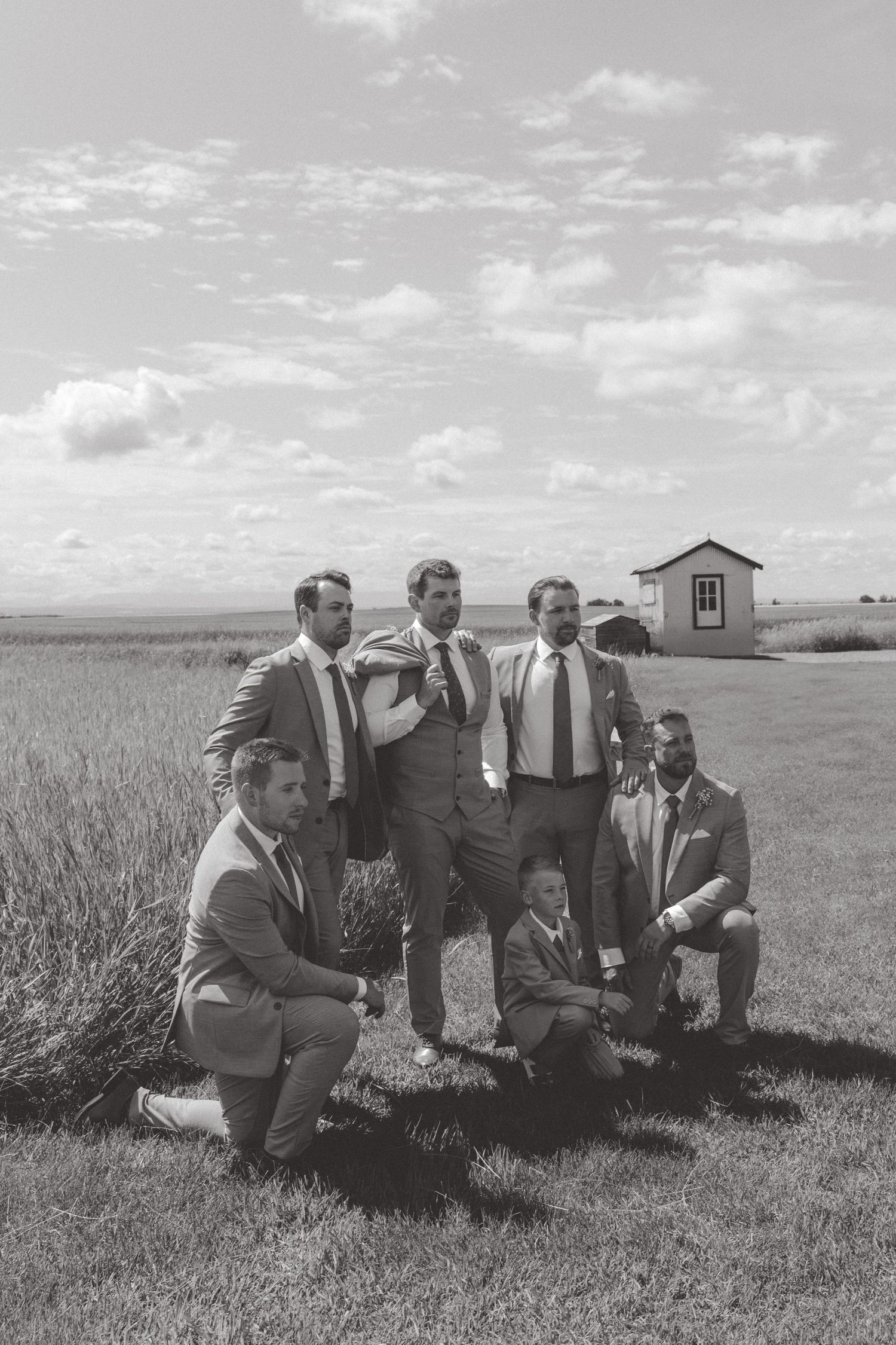 Black and white photo of groomsmen relaxing on grass during outdoor wedding.