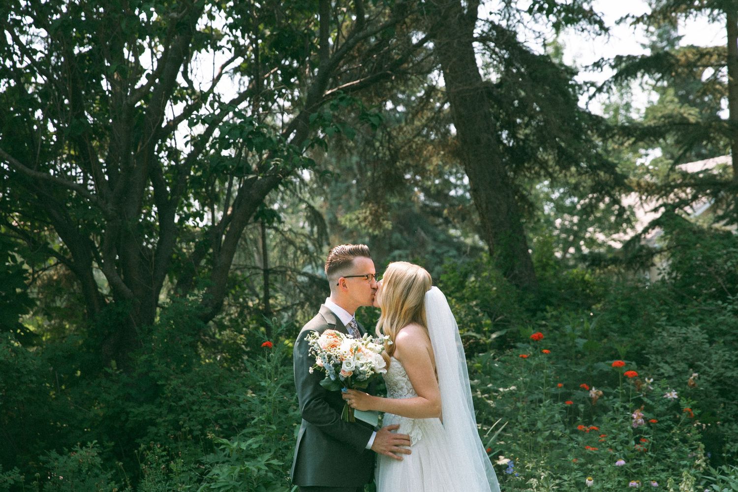 A romantic wedding embrace captured in a garden setting with towering trees.