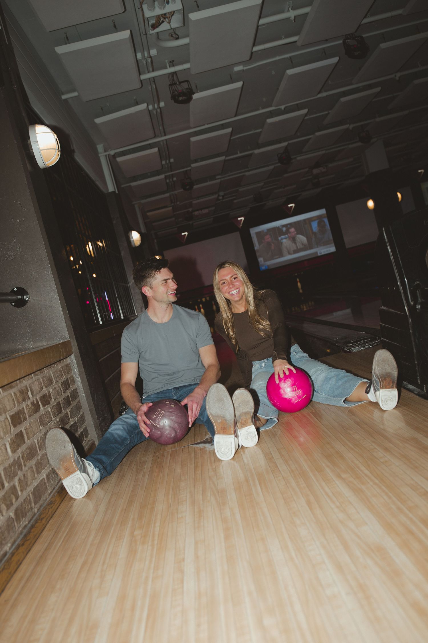 A smiling couple sits on a bowling alley lane holding pink bowling balls, enjoying a fun date night.