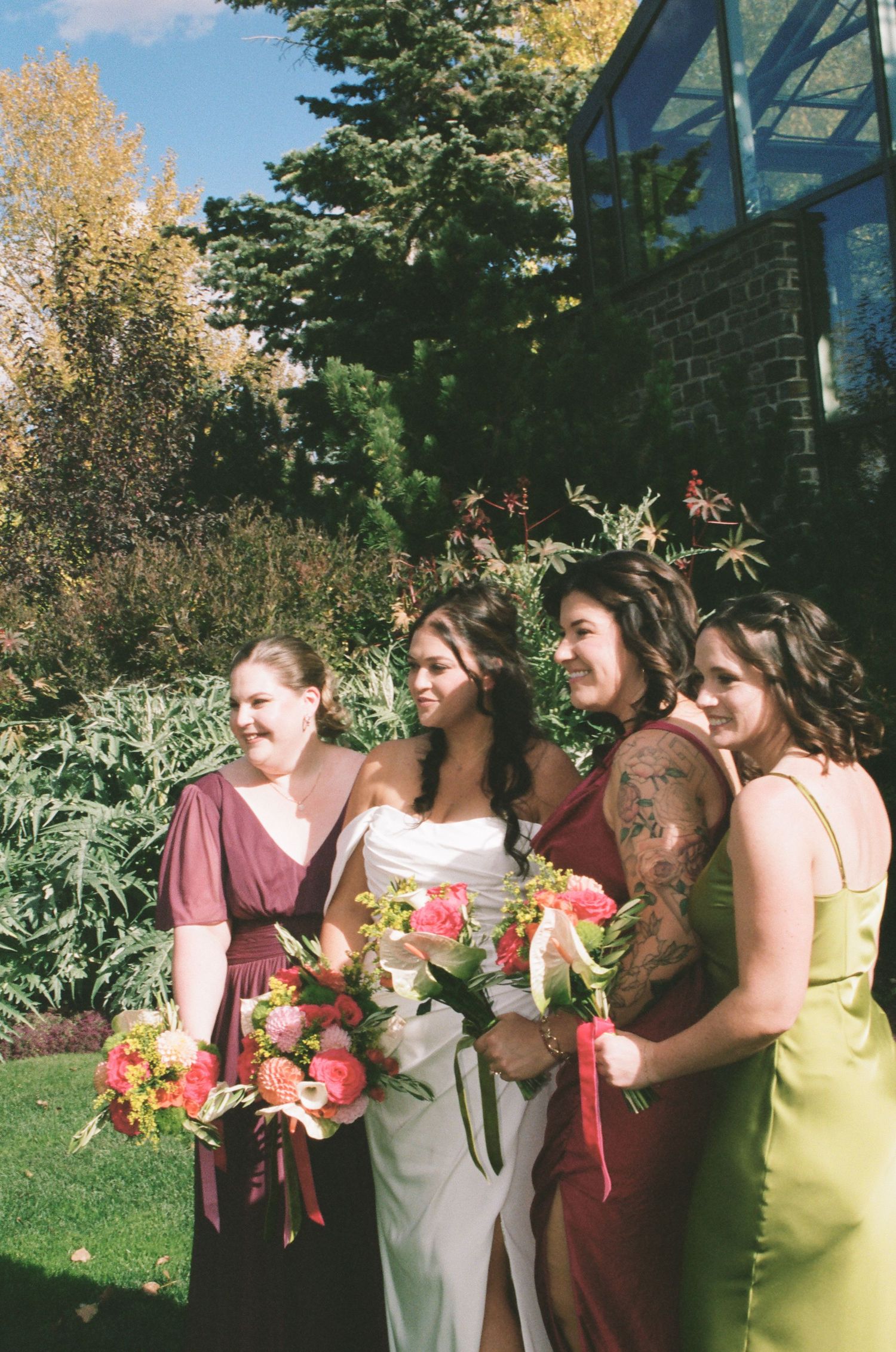 A bride in white with three bridesmaids in colorful dresses holding bouquets outdoors.