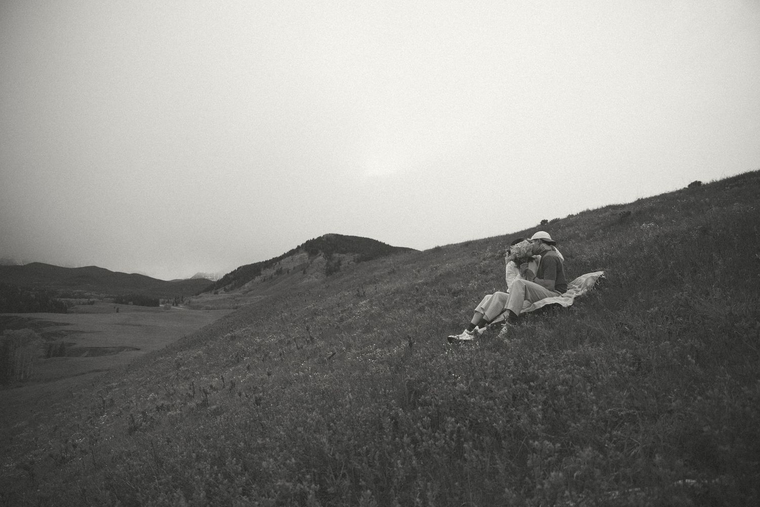 Black and white photo of a couple sitting on a hillside overlooking a scenic landscape.