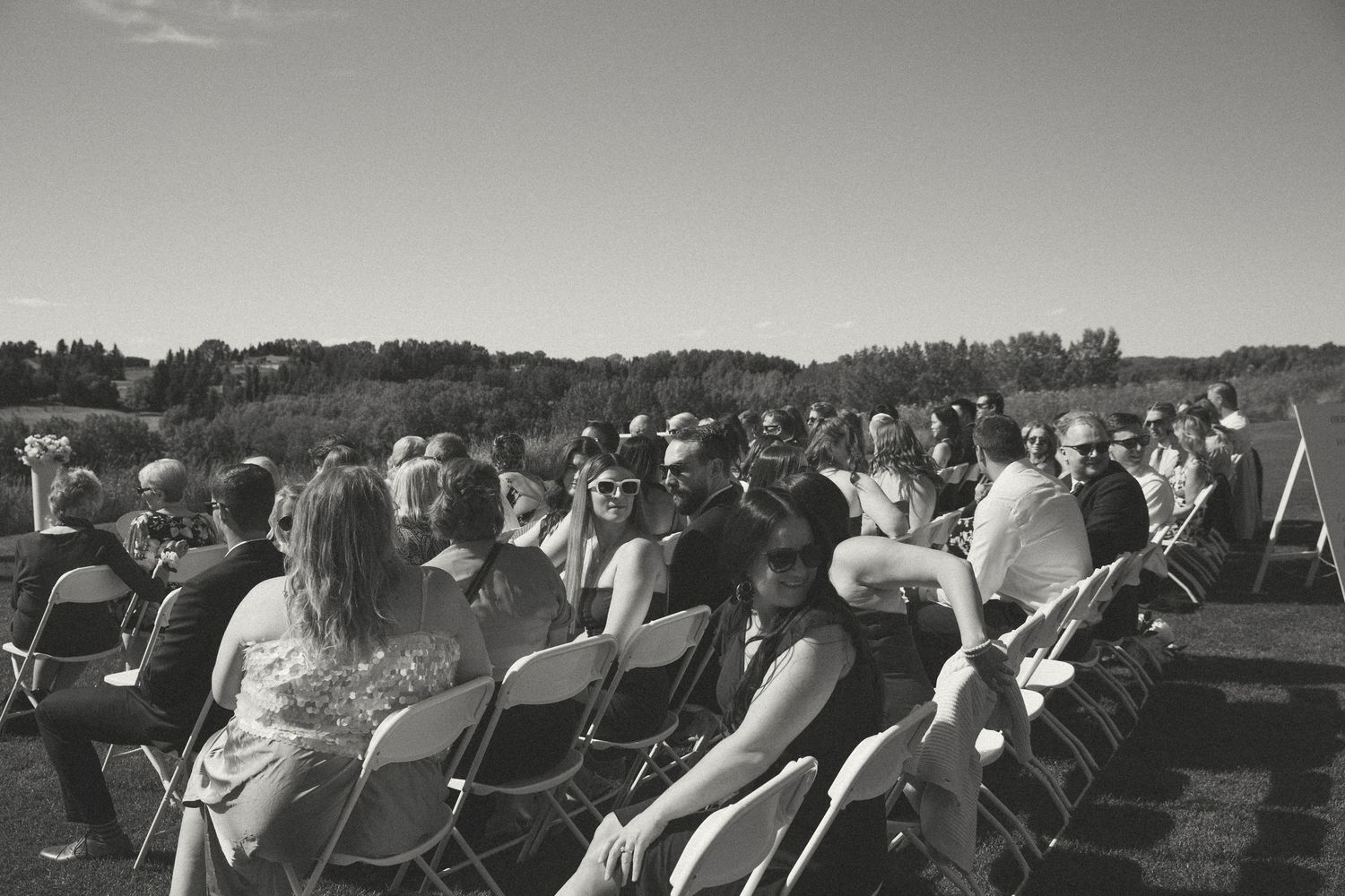 Black and white photo of wedding guests seated in white chairs at outdoor ceremony.