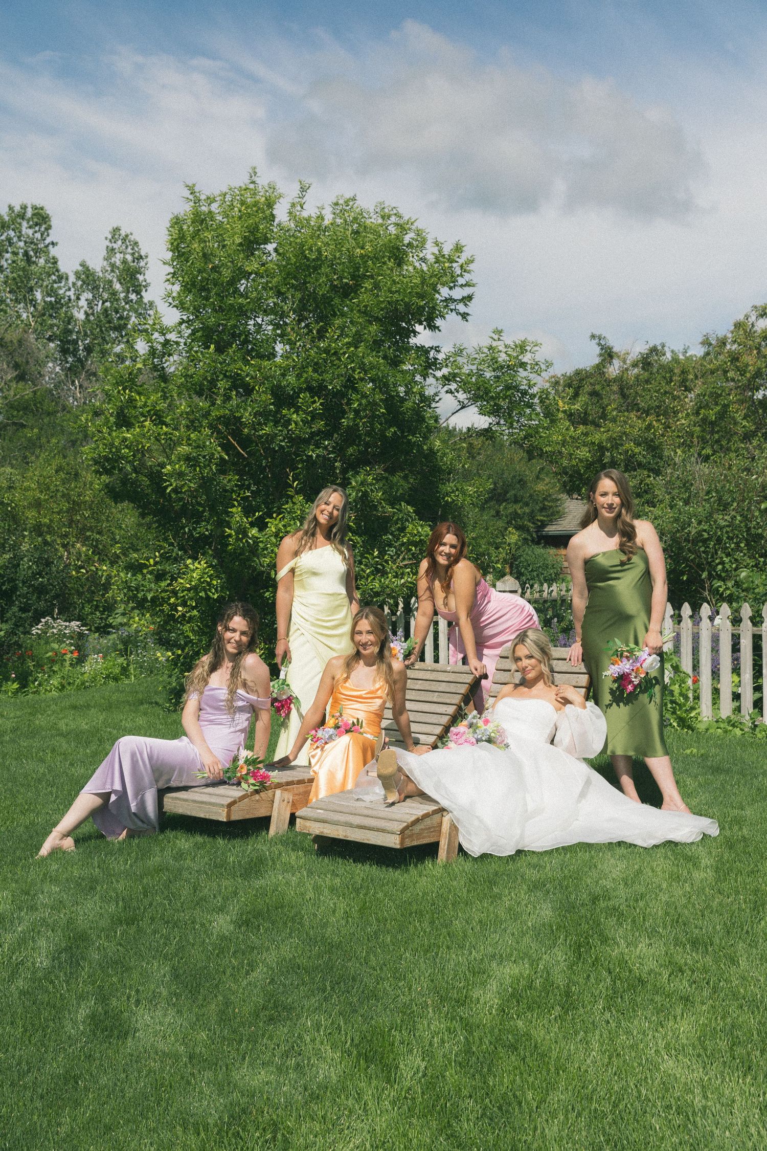 Wedding party relaxes on wooden benches in a sunny garden setting.