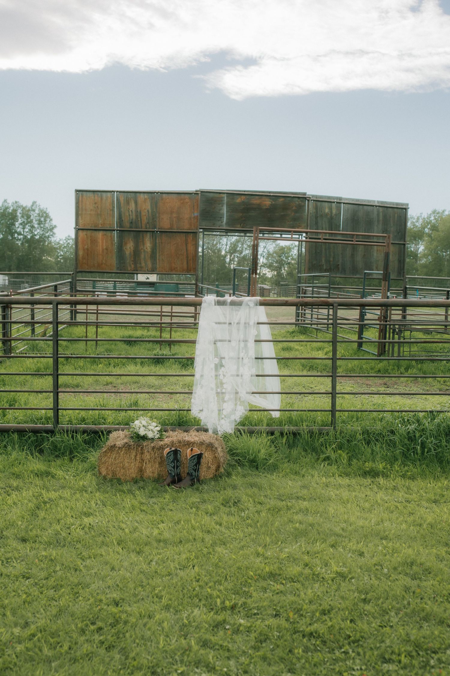 Wedding dress hanging on rustic metal farm fence over hay bale in green field.